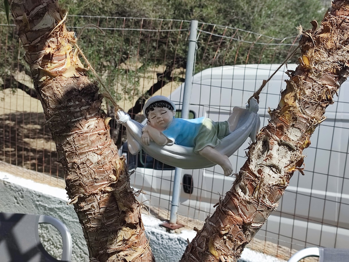 A decorative figurine is shown resting in a small hammock, suspended between two tree trunks. The figure is dressed in a light blue outfit, conveying a sense of relaxation. Behind the figurine, a fence and van are partially visible, integrating the garden setting.