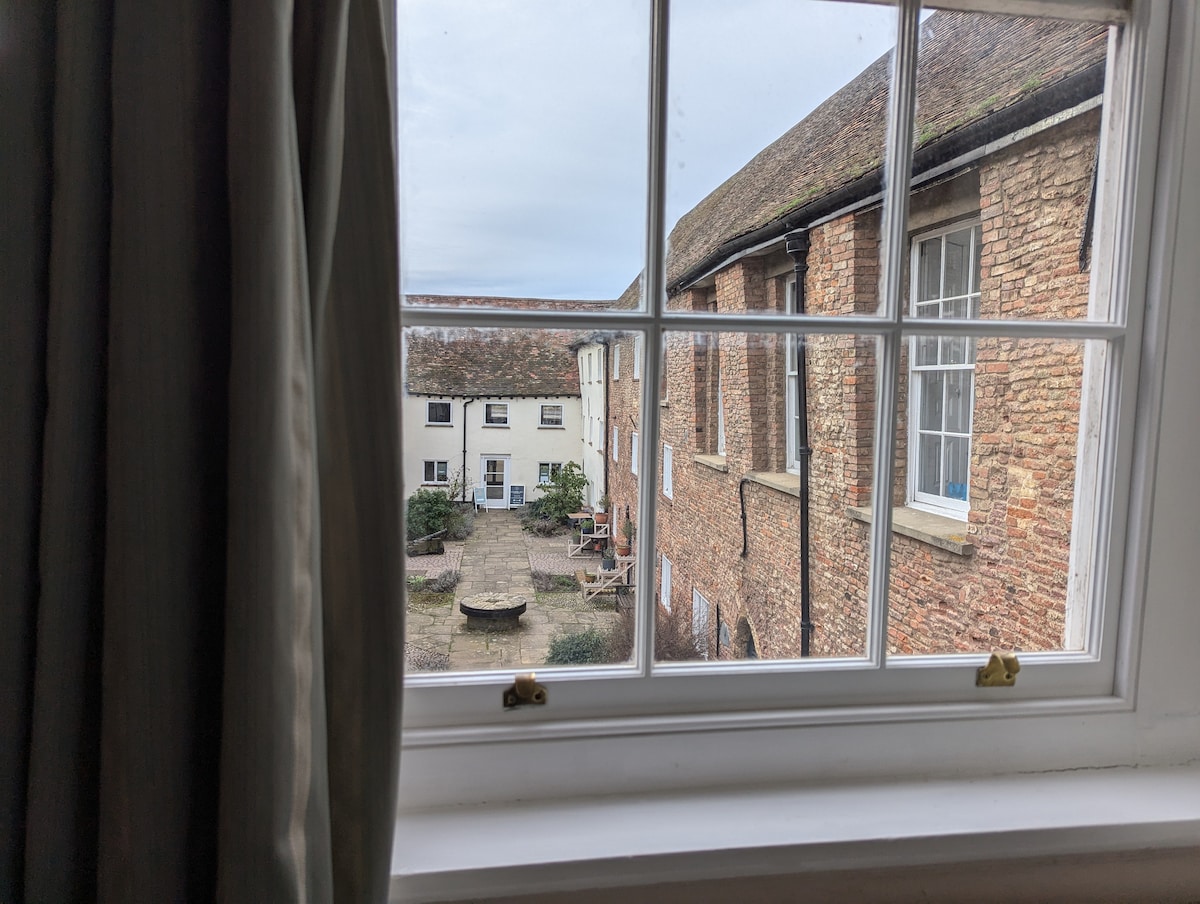 A view from a window showcases the courtyard of Hanse House, featuring a circular stone seating area surrounded by greenery. The historic brick buildings frame the scene, highlighting the charm of the cobbled streets and inviting ambiance of the surroundings.