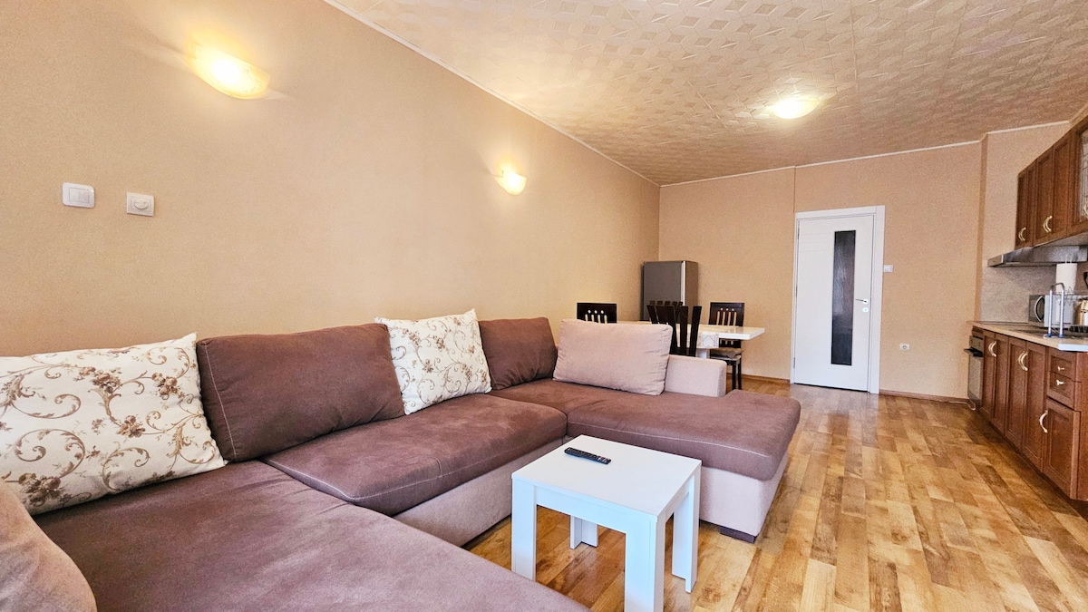 A spacious living area is highlighted by a large sectional sofa adorned with decorative pillows. A small coffee table is positioned in front of the sofa. Light wooden flooring complements the cream-colored walls, while a dining table with chairs is visible in the background.