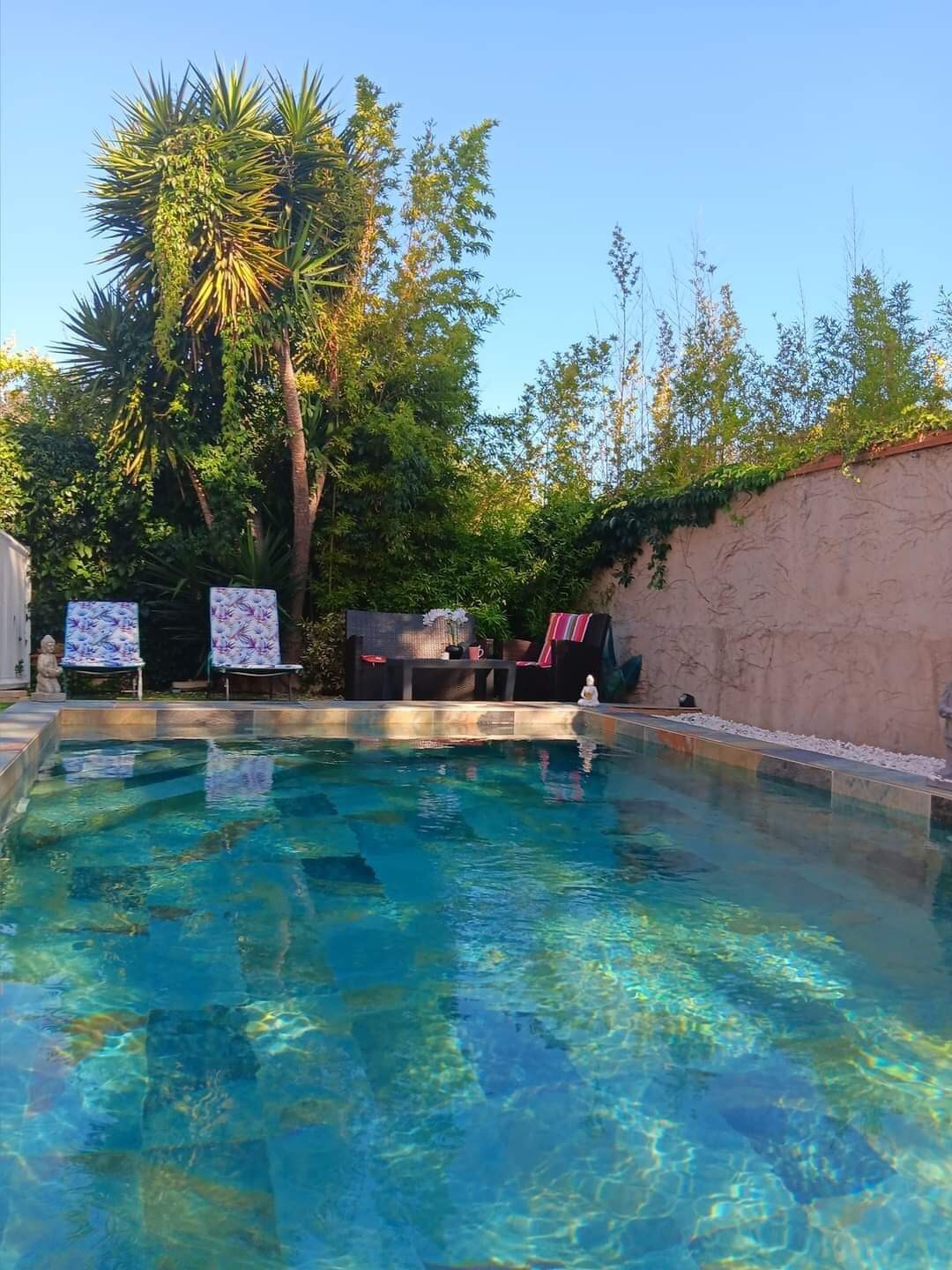 A serene pool area is framed by lush greenery, featuring clear blue water that reflects the sunlight. Two colorful lounge chairs are positioned nearby, alongside a shaded seating area, inviting relaxation in a peaceful setting.