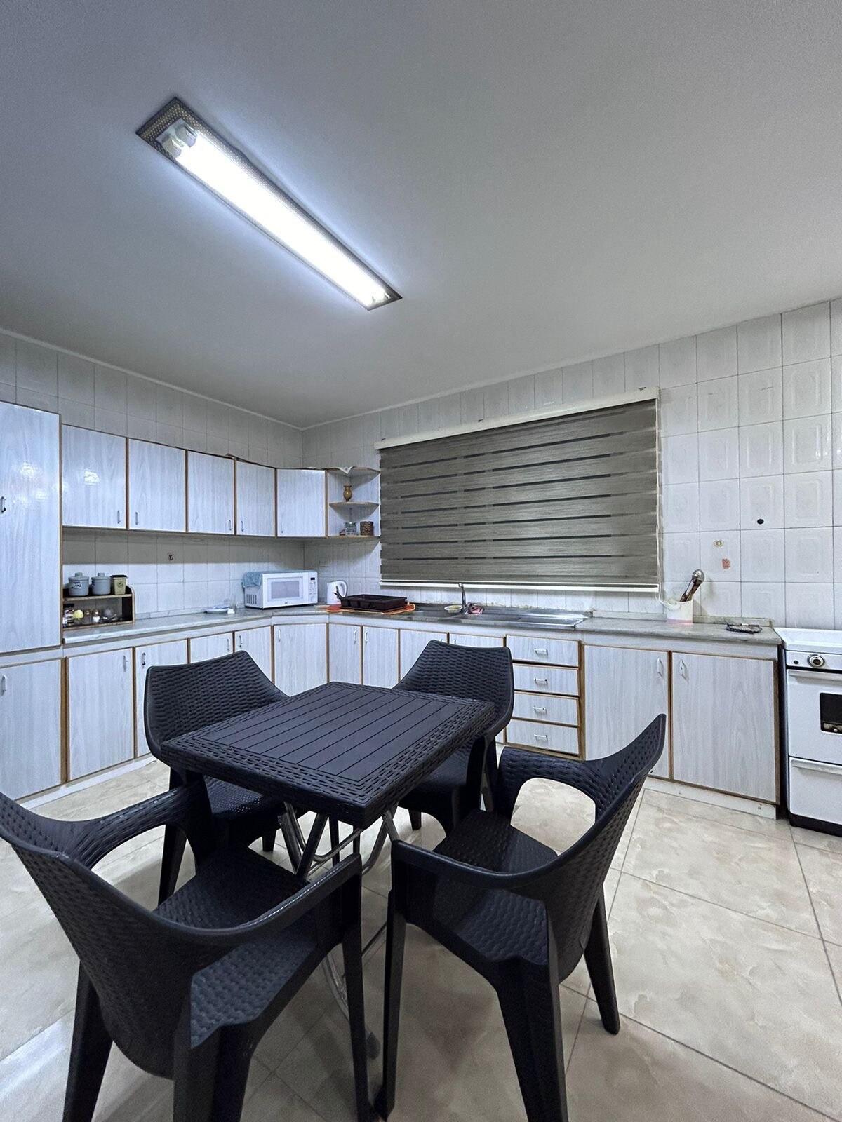 A functional kitchen area is presented with light grey cabinetry and a compact black dining table surrounded by four black chairs. Essential appliances, including a microwave and stove, are visible, along with a window that allows natural light to illuminate the space.