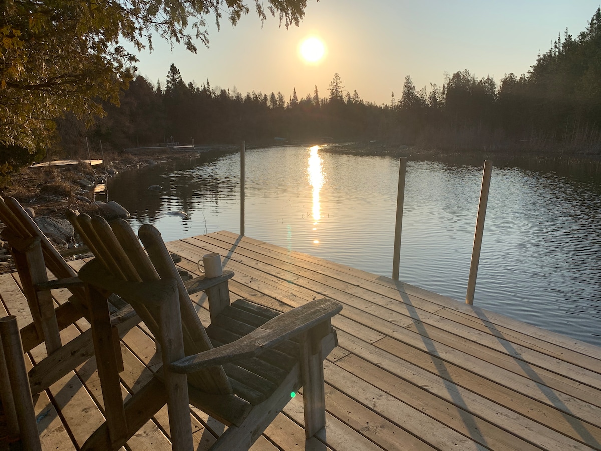 A wooden dock extends over calm waters, with a pair of rustic chairs positioned invitingly. The setting sun reflects shimmering light on the surface of the water, surrounded by trees in the background, creating a serene atmosphere.