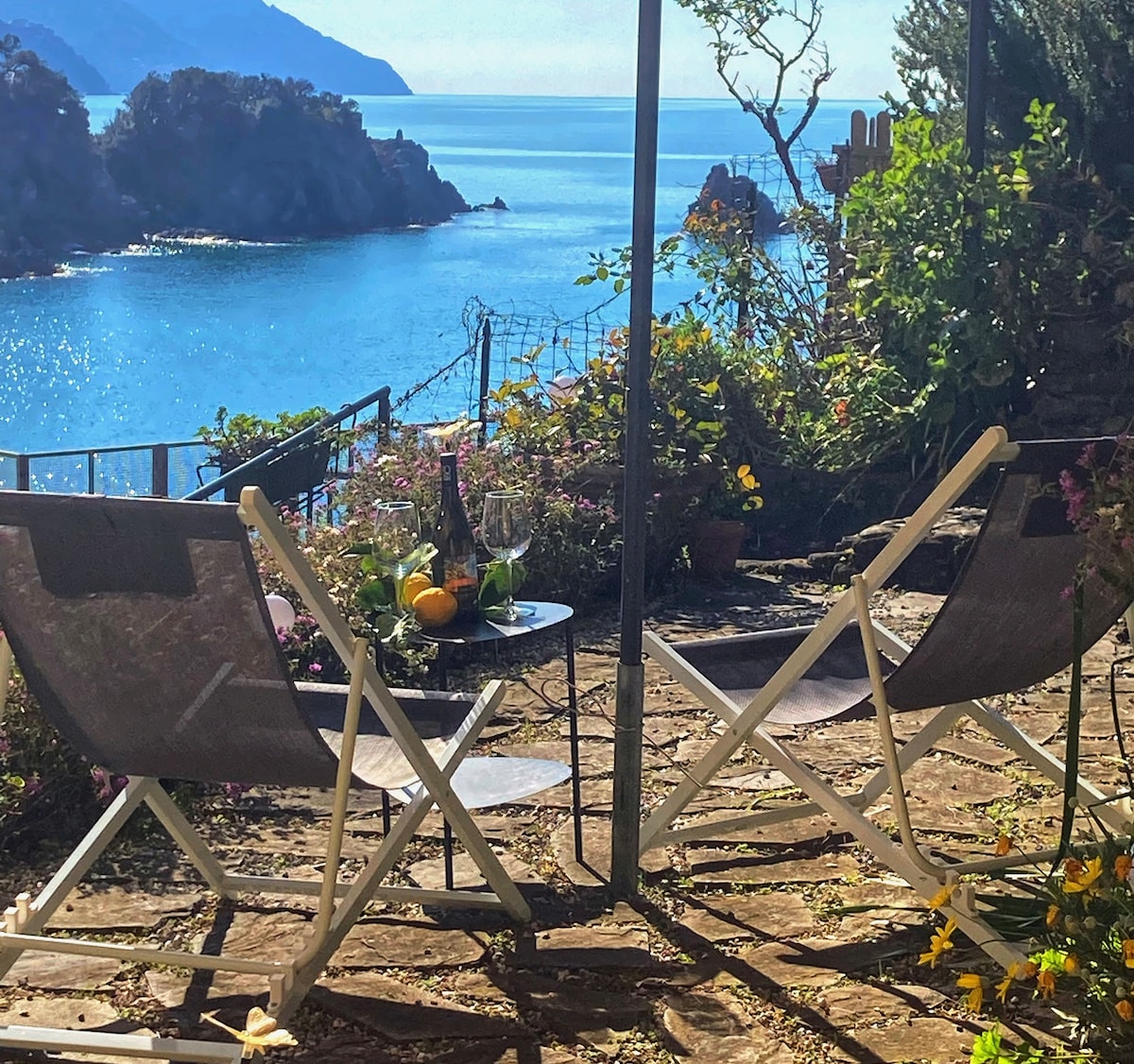 A serene outdoor space features two folding chairs facing a panoramic sea view, with vibrant flowers and greenery surrounding the area. A small table is set with a bottle of wine and fresh fruits, providing an inviting spot for relaxation amidst the natural landscape.
