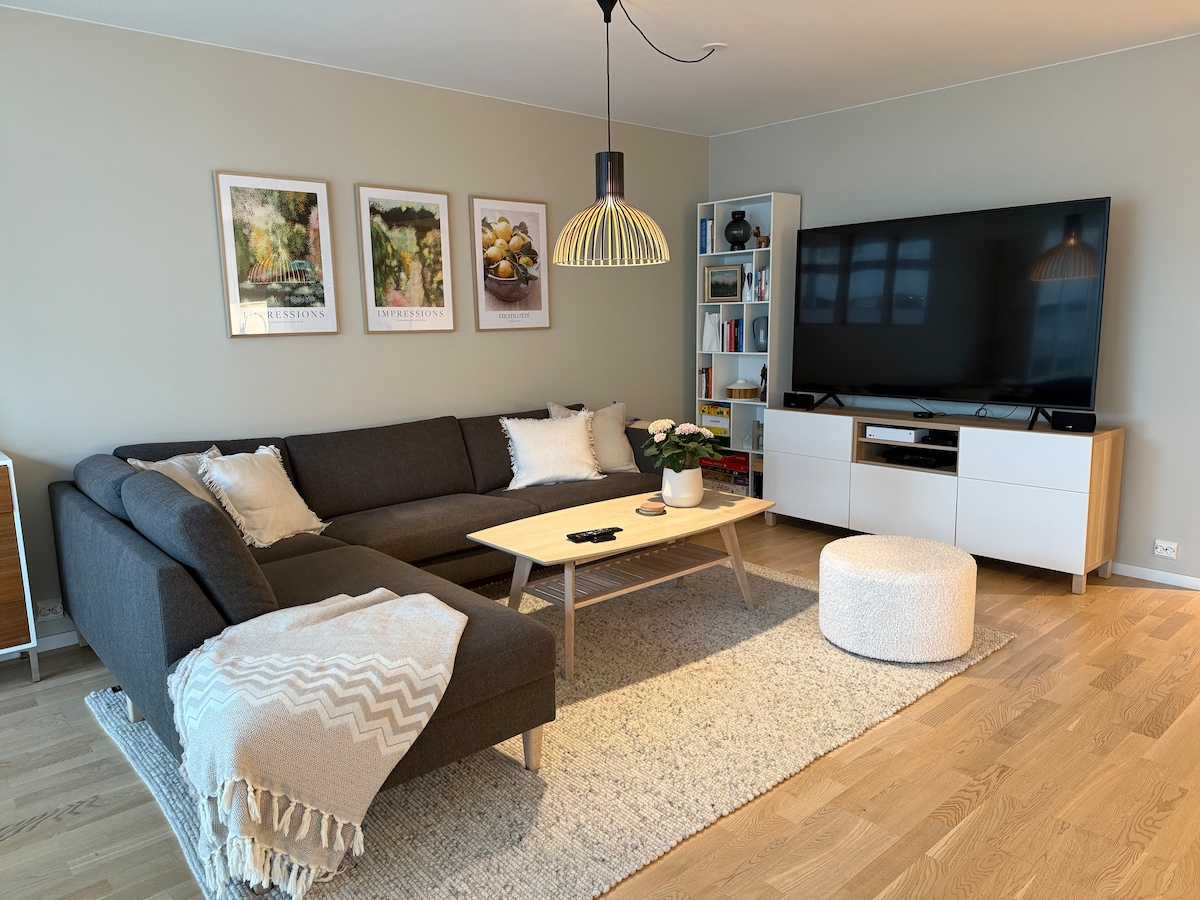 A spacious living room is presented with a comfortable gray sectional sofa, adorned with decorative cushions and a soft blanket. A light wooden coffee table is centered on a textured rug, accompanied by a rounded ottoman. Natural light is welcomed by large windows, and a large TV is positioned on a sleek white media unit.