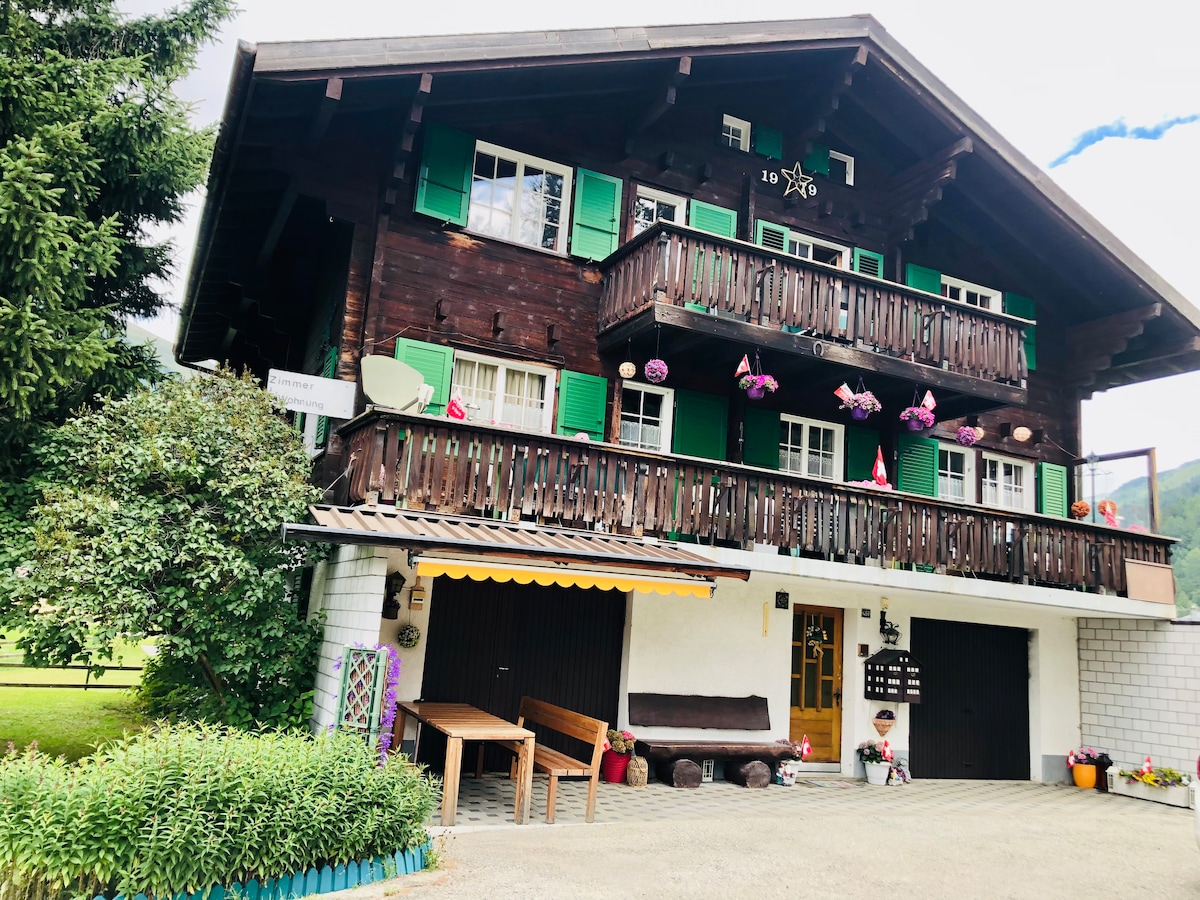 A traditional chalet is presented, featuring a wooden exterior with green shutters and flower boxes. Two balconies adorned with potted plants enhance the charming facade. A spacious seating area is visible in front of the entrance, surrounded by lush greenery and a gravel driveway.