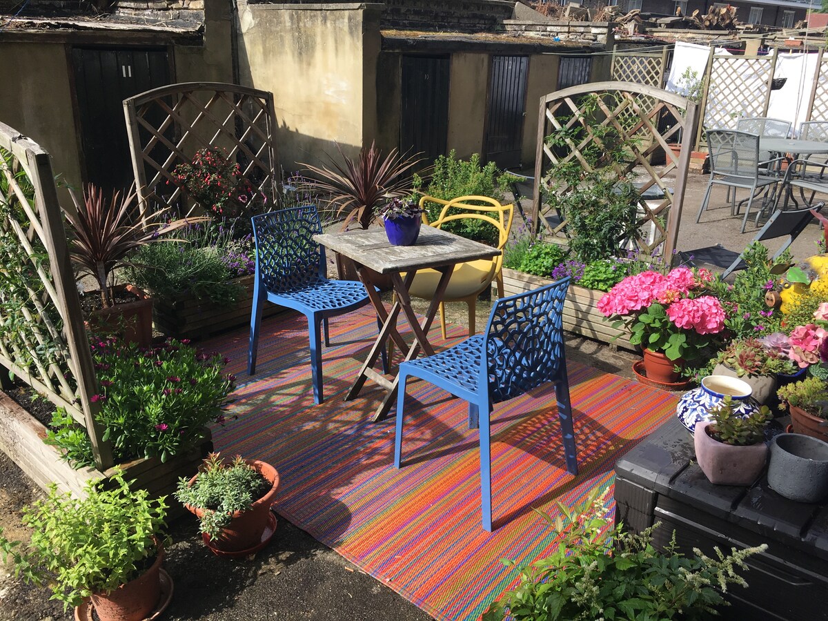 A vibrant garden space is showcased, featuring a small table with two colorful chairs, surrounded by lush greenery and blooming flowers. Decorative planters add character, and a striped outdoor rug anchors the seating area, providing a cheerful and inviting atmosphere for outdoor dining.