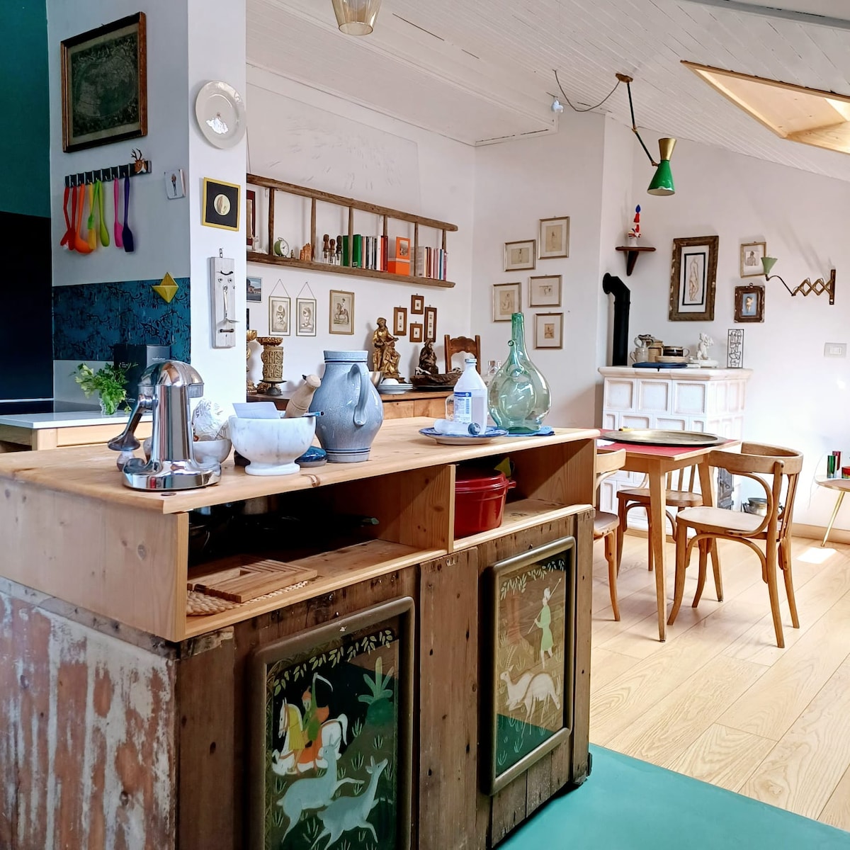 An open kitchen area features rustic wooden counters with decorative elements displayed. Artworks and framed pictures adorn the walls, while a dining table with chairs is positioned nearby. Natural light enters through a skylight, creating a warm and inviting space.