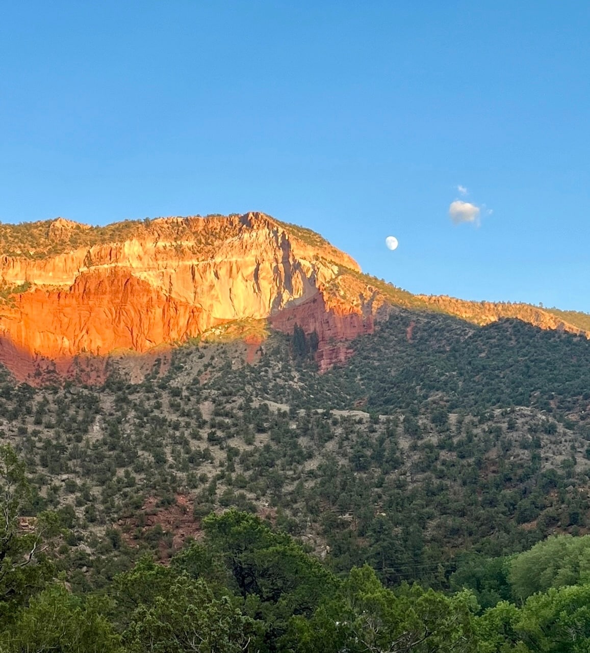 A stunning mountain rises, displaying rich hues of red and orange as sunlight casts warm tones on the rocky face. The sky features a soft blue backdrop, with a moon visible, contributing to the serene landscape surrounded by lush greenery.