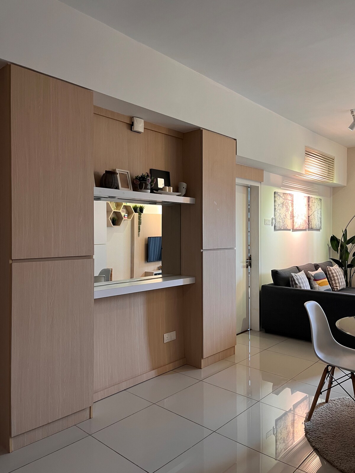 A minimalist living space is highlighted by wooden cabinetry that encloses a reflective mirror and decorative plants. Bright tiles extend across the floor, and a cozy seating arrangement with a black sofa is visible in the background, complemented by soft lighting and wall art.