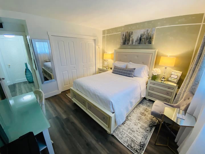 Útulná spálňa s manželskou posteľou Queen, 2 nočnými stolíkmi s lampami, šatníkom, vešiakmi, žehliacou doskou, minichladničkou, mikrovlnnou rúrou, stolom so stoličkou, kávovarom, televízorom, zrkadlom a 2 plážovými stoličkami.