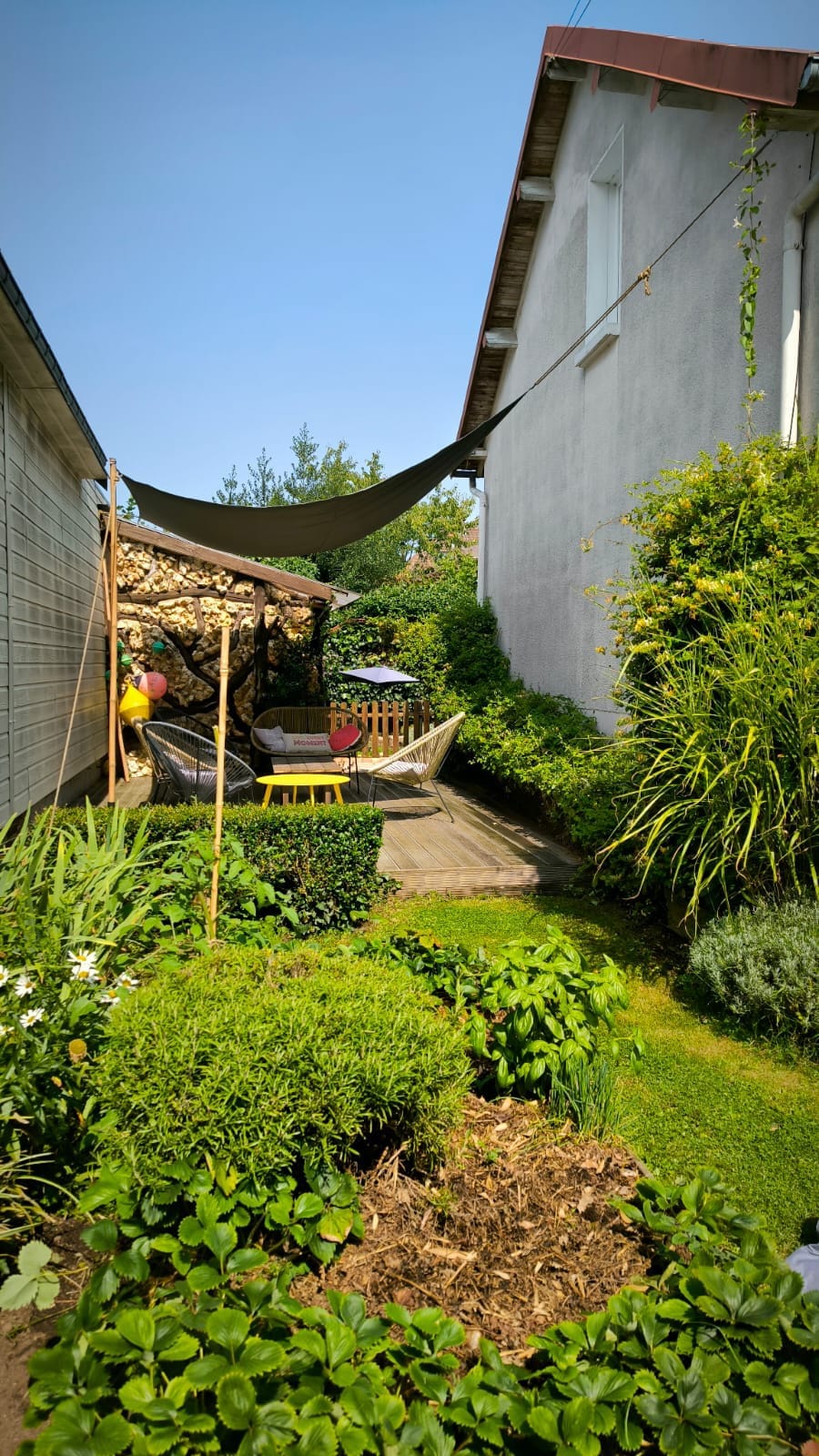 A lush garden pathway is surrounded by greenery, featuring vibrant plants and flowers. A shaded seating area with a hammock and a small table is nestled under a canopy. Wooden decking provides a relaxing space, enhancing the outdoor ambiance.