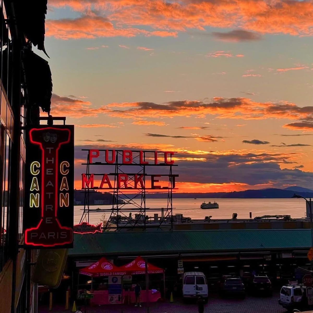 A vibrant sunset casts hues of orange and pink across the sky, with the iconic Public Market sign clearly visible. The ocean reflects the warm colors, creating a serene backdrop. Silhouettes of boats can be seen in the distance, adding to the atmosphere.