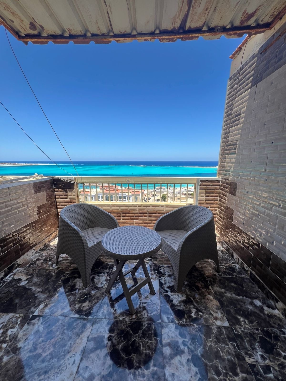 A small balcony offers two woven chairs and a round table, overlooking a vibrant blue sea. The smooth, marble flooring is complemented by a backdrop of gray tiled walls, enhancing the coastal view.