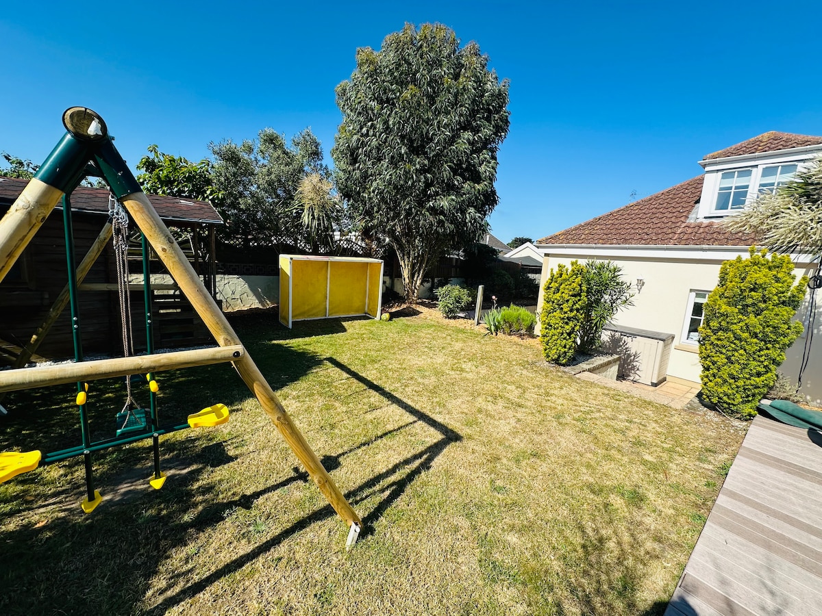 The large garden features a grassy area, adorned with a swing set and a yellow play structure. Lush greenery surrounds the space, and a light blue sky enhances the openness of the area. Patio space is visible on one side, suggesting a comfortable outdoor setting.
