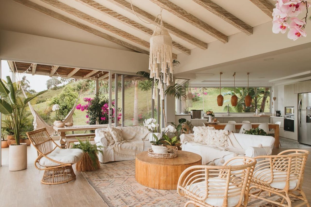 A welcoming living area features large glass doors that open to a lush outdoor landscape. Natural light fills the space, highlighting a round wooden coffee table surrounded by comfortable seating. Elegant decor, including plants and wall hangings, add to the relaxed ambiance.