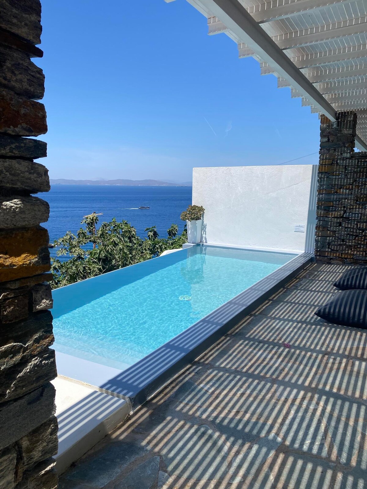 A clear view of a private infinity pool is presented, reflecting the vibrant blue sea beyond. Sunlight filters through a trellis, casting striped shadows on the deck. Stone walls frame the scene, enhancing the connection between indoor and outdoor spaces.