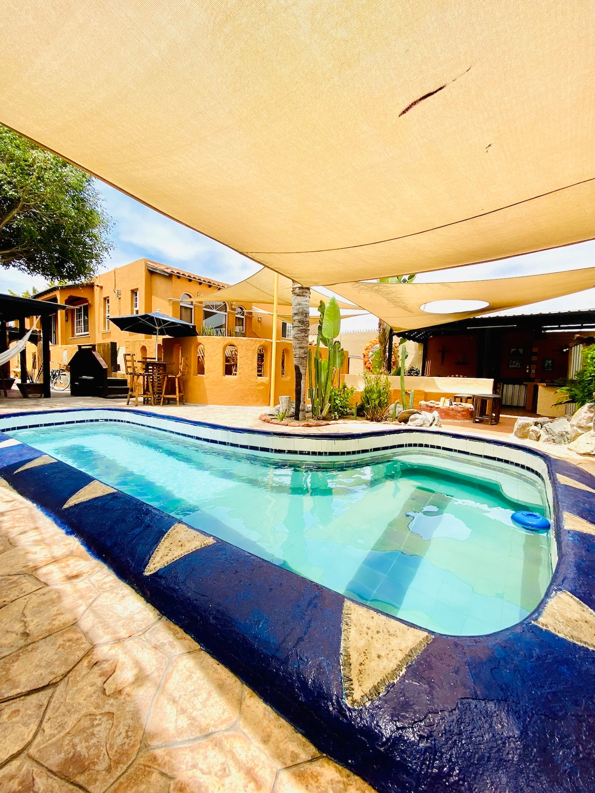 An inviting L-shaped private pool is surrounded by shaded areas under a fabric canopy. Tropical plants and natural stone decor frame the pool, while adjacent spaces include outdoor seating areas with tables and umbrellas, providing a relaxed atmosphere for enjoying the outdoors.