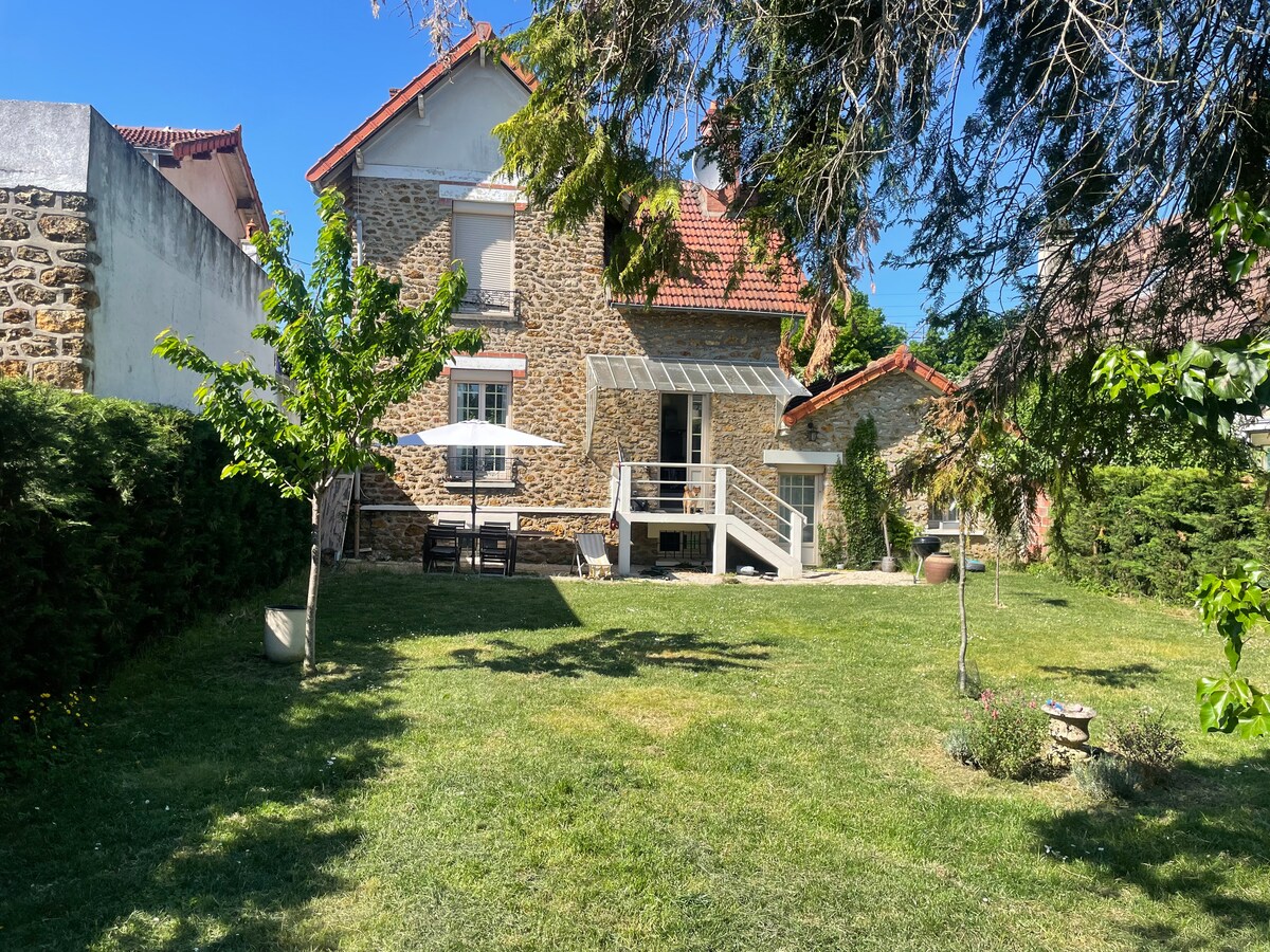 The exterior of the cozy two-room apartment is highlighted, showcasing a spacious shared garden with lush green grass. A patio area features an umbrella and table, providing a charming spot to relax outdoors. The stone structure of the apartment complements the serene residential surroundings.