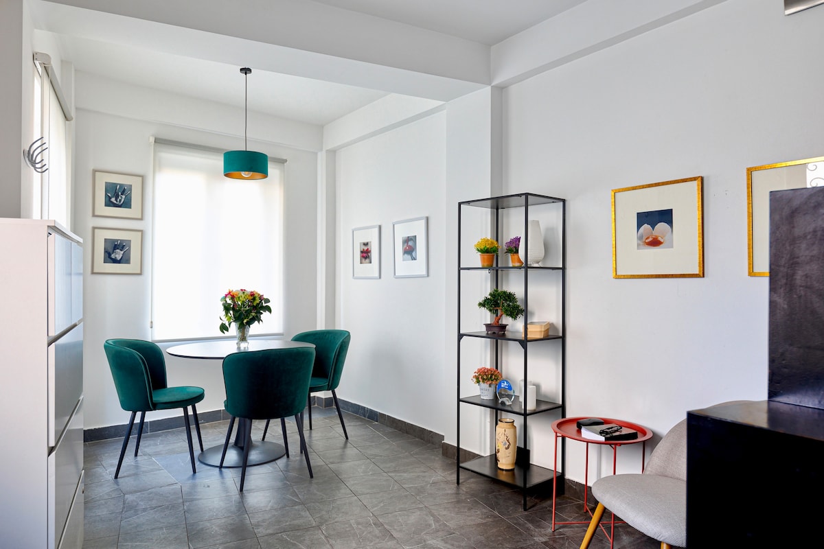 A dining area is presented with a circular table surrounded by four teal chairs. Natural light filters through a window, highlighting the room's neutral tones. A tall, black shelf displays decorative items and plants, while framed artwork adorns the white walls.