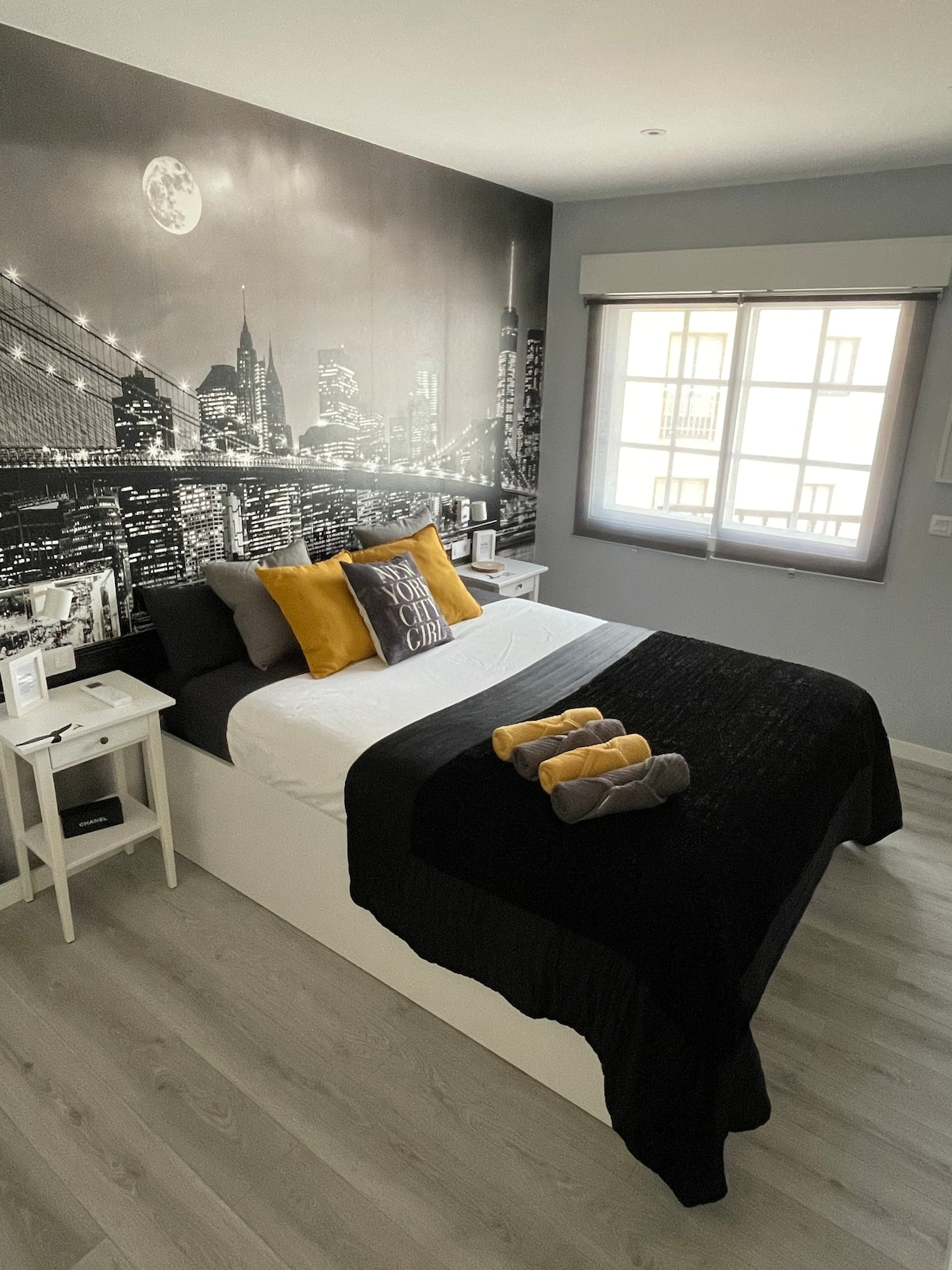 A modern bedroom features a spacious bed with a black coverlet and decorative pillows in shades of yellow and gray. A large cityscape mural adorns the wall behind the bed. A window allows natural light to fill the room, complemented by a light-colored wooden floor.