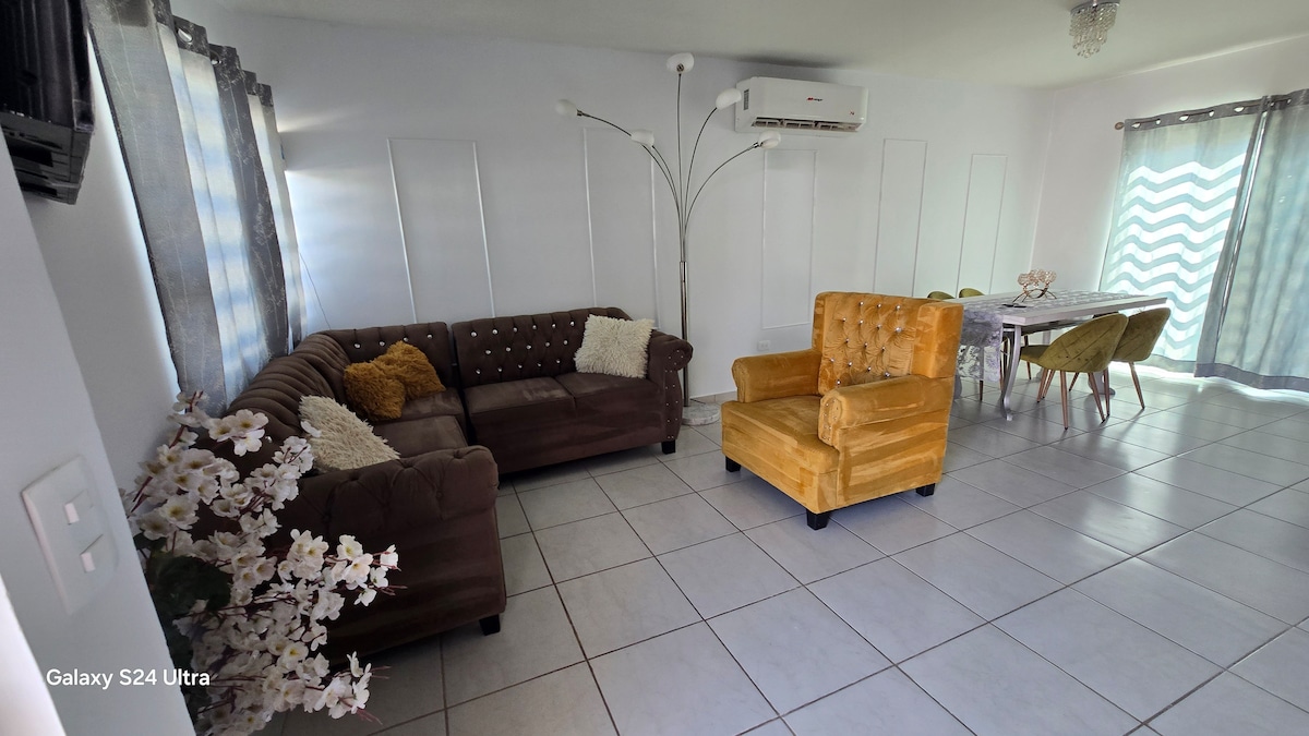 A living area features two brown sofas and a golden armchair arranged around a glass-topped table. Natural light streams through sheer curtains, highlighting the ceramic tiled floor. A floor lamp stands in the corner, and air conditioning is visible on the wall.