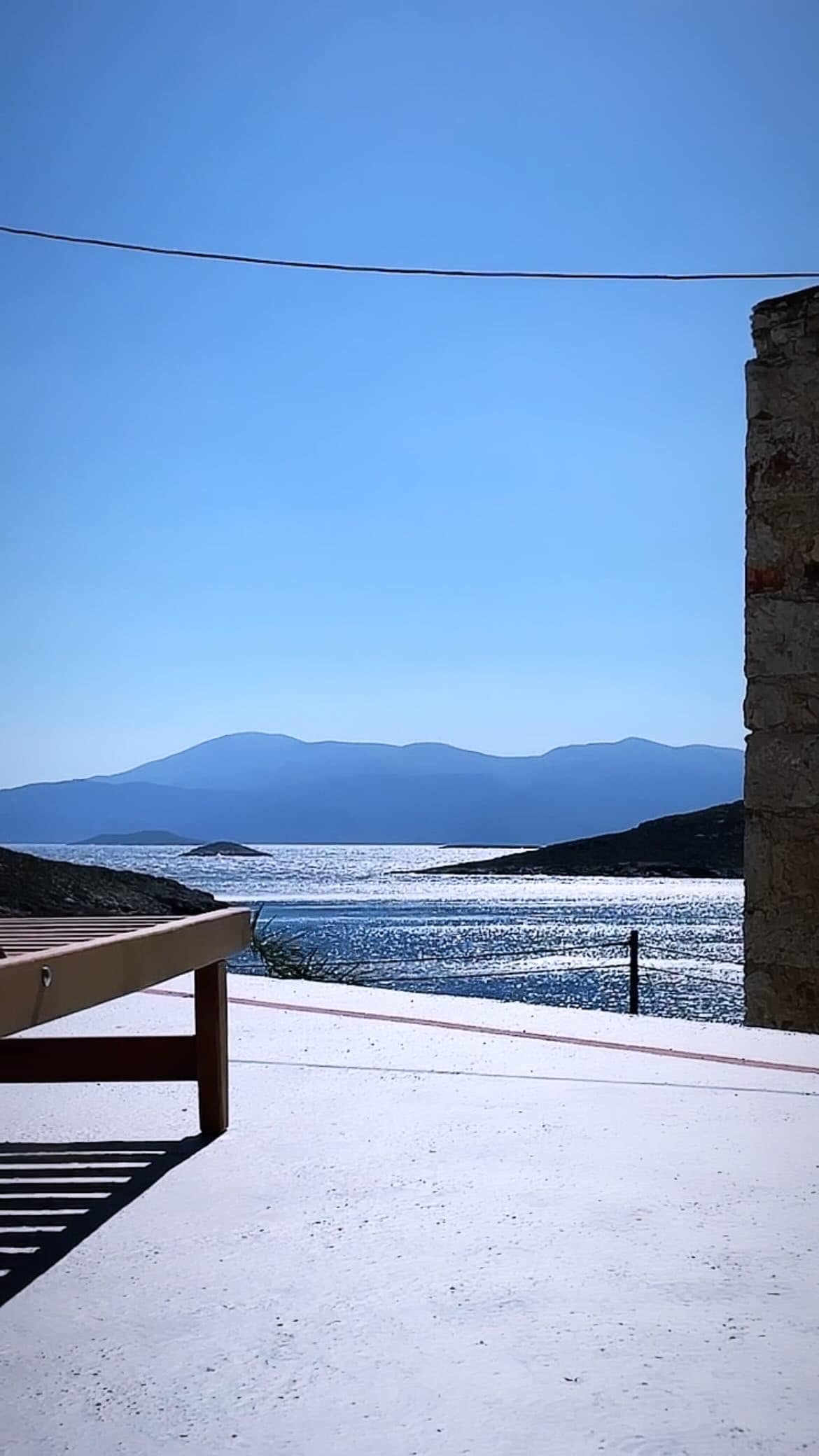 A calming view of the ocean is framed by a stone wall, with distant mountains visible under a clear blue sky. The water reflects sunlight, creating a serene atmosphere. A wooden lounge chair is positioned on the white terrace, inviting relaxation.