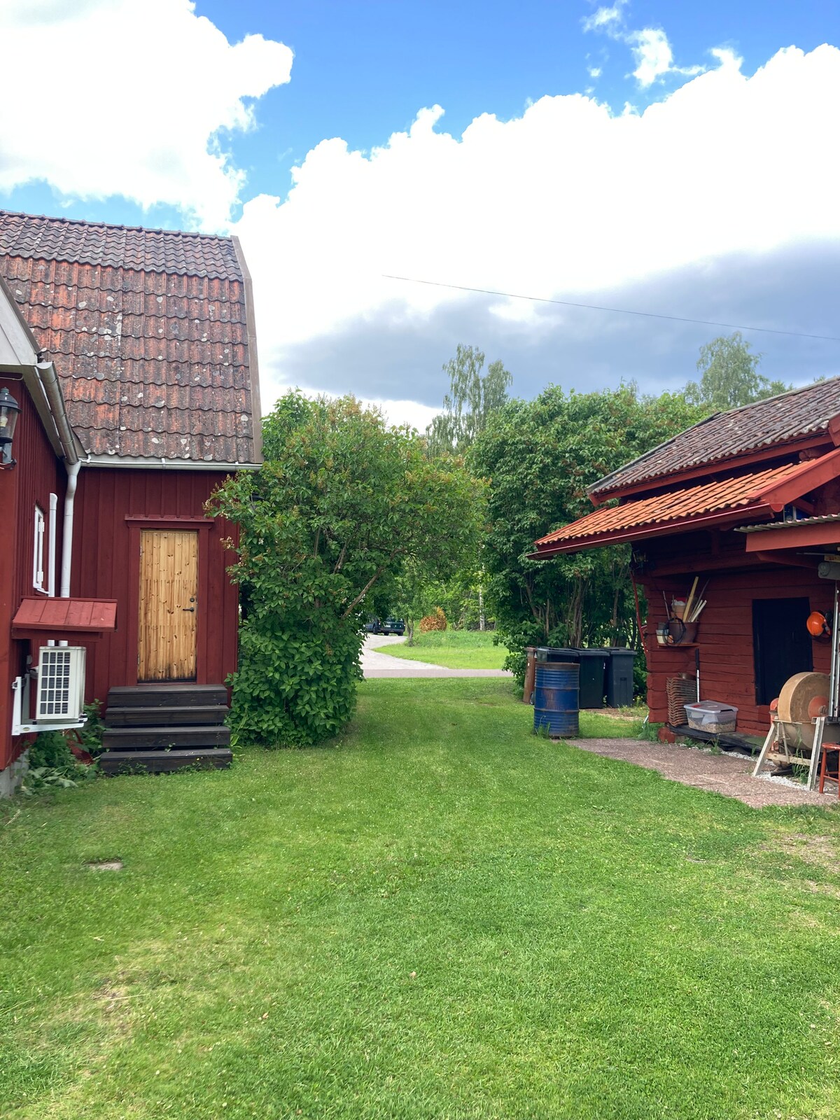 An inviting outdoor space is depicted, featuring a grassy path that runs between two traditional red wooden structures. Lush green bushes and trees provide a natural boundary, with a clear sky visible above, offering a serene setting for relaxation.
