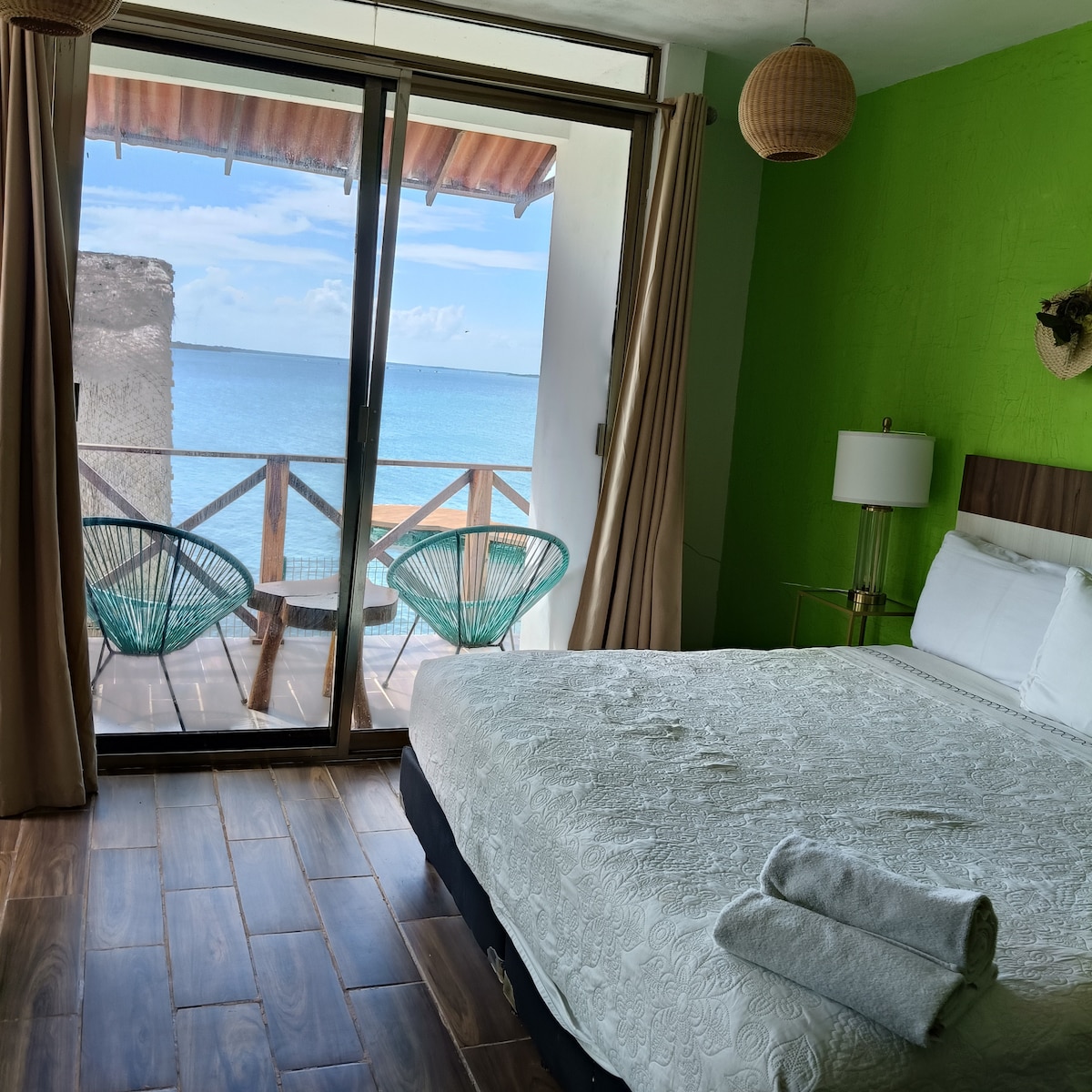 A spacious sleeping area is featured, showcasing a kingsize bed with soft linens. Large glass doors lead to a private balcony with views of the lagoon. The wall is painted in a vibrant green, complemented by woven light fixtures and wooden accents.