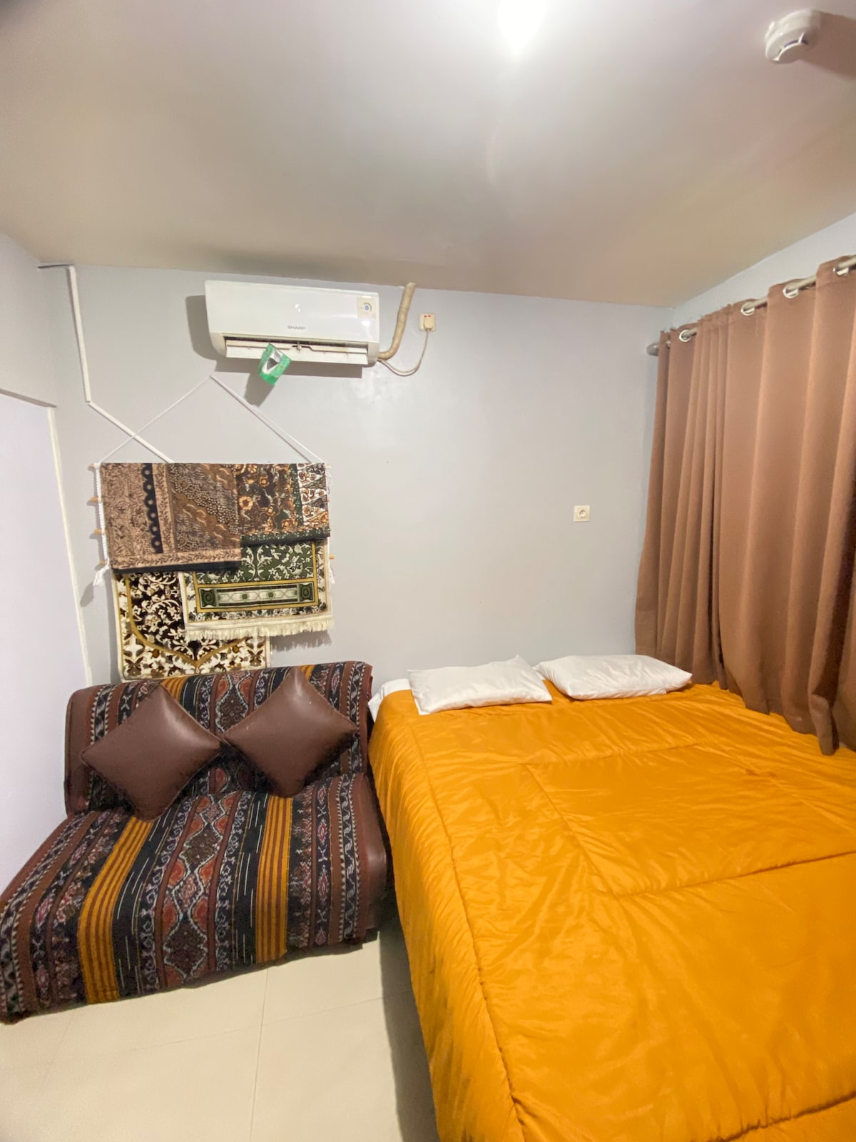 A compact room features a spacious bed covered in a warm orange duvet, paired with two decorative pillows. A small couch with patterned fabric and two brown cushions is located beside the bed. Light curtains frame the window, and an air conditioning unit is mounted on the wall.