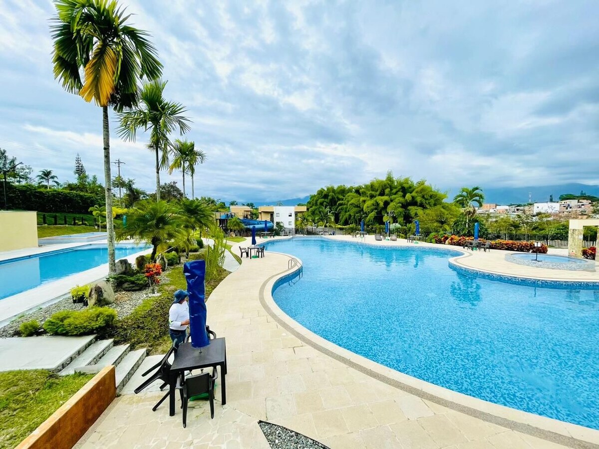 A large, inviting pool area features multiple swimming pools surrounded by well-maintained gardens. Palm trees and vibrant landscaping create a serene environment, while lounge chairs and tables are positioned for relaxation. The gentle curves of the pool edges enhance the tranquil atmosphere.
