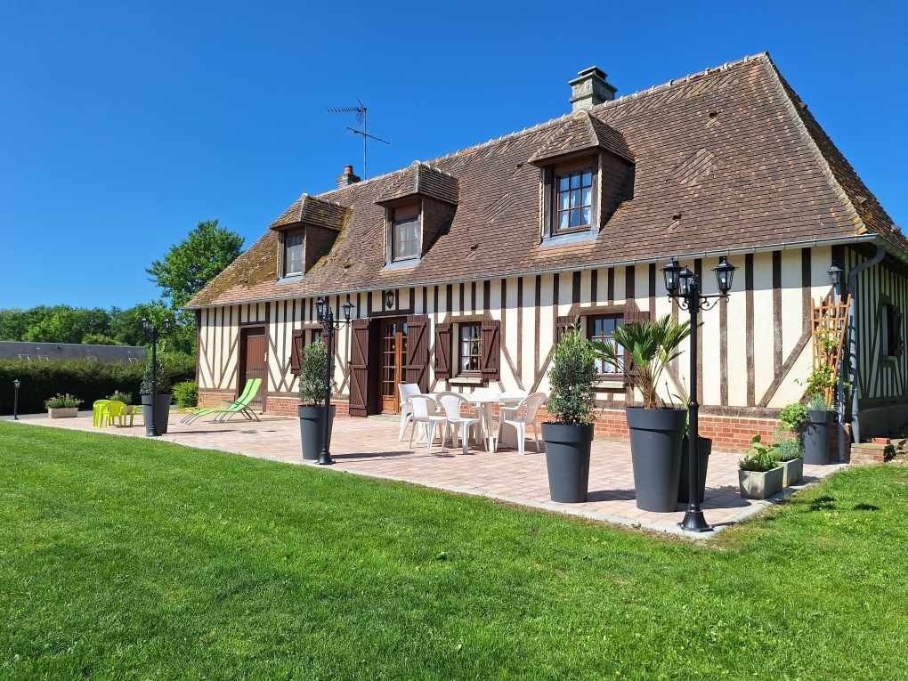 A charming countryside house displays traditional half-timbered architecture under a clear blue sky. The outdoor terrace is furnished with a dining table and chairs, surrounded by potted plants. Lush green grass extends in front of the house, enhancing the serene outdoor setting.