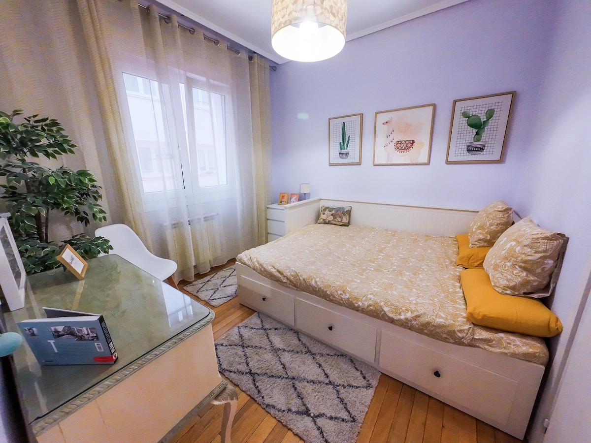 A cozy bedroom features a spacious bed with yellow accent pillows and a light-colored blanket. On the walls, three framed botanical prints add a touch of nature. A small desk and chair are positioned near a window draped with sheer curtains, allowing natural light to brighten the room.