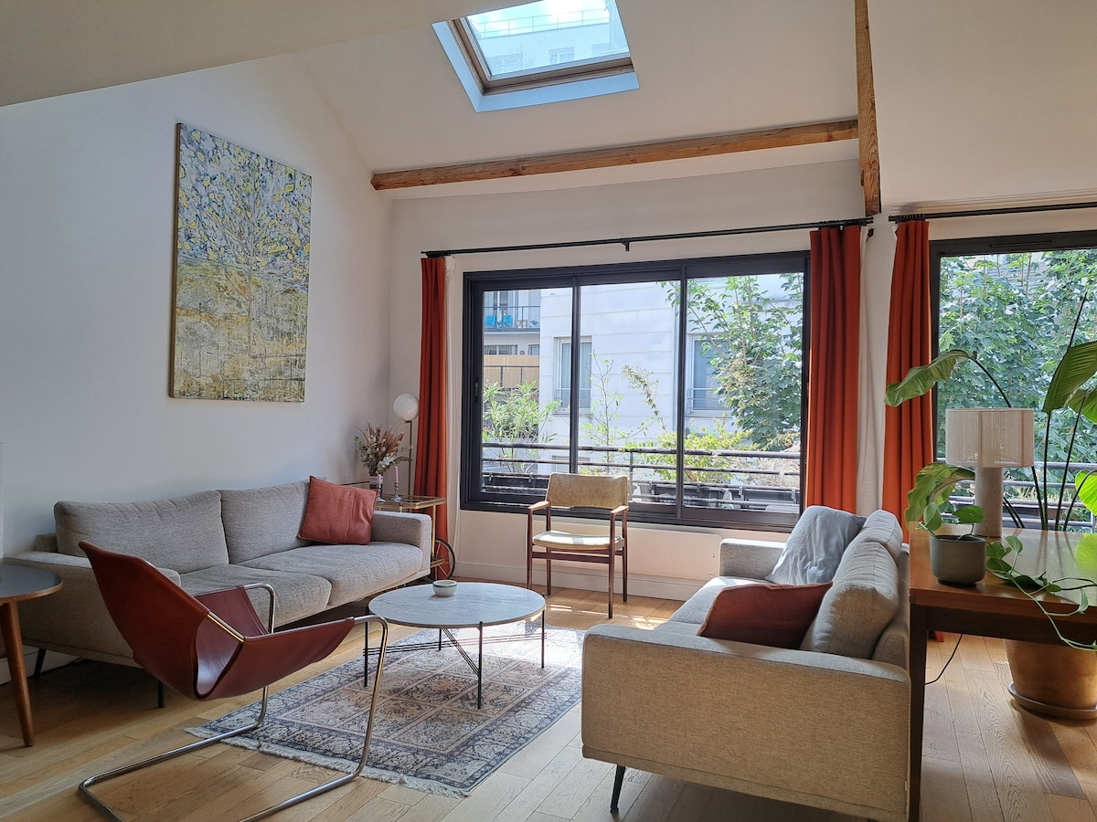A spacious living area features two sofas, a coffee table, and a chair, all arranged to encourage conversation. Large windows with curtains allow natural light to fill the room, while a Velux window above enhances brightness. A painting and a potted plant add subtle decor.