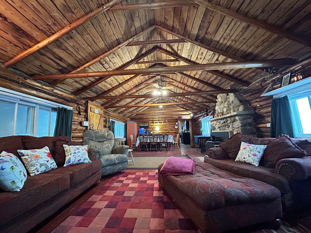A spacious cabin interior is highlighted by wooden beams and a high ceiling. Comfortable seating areas are arranged on a patterned rug, with decorative pillows on a sofa. A dining table is visible in the background, accompanied by a stone fireplace that adds rustic charm.