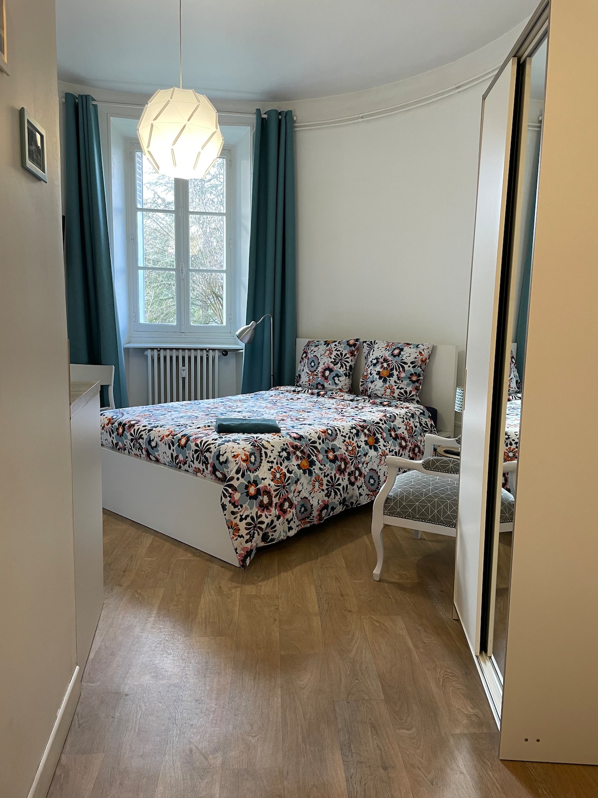 A bright sleeping area features a double bed adorned with a colorful patterned duvet. A bedside table holds a small lamp, and a full-length mirror reflects the space. Light filters through a large window dressed in turquoise curtains, enhancing the relaxing atmosphere.