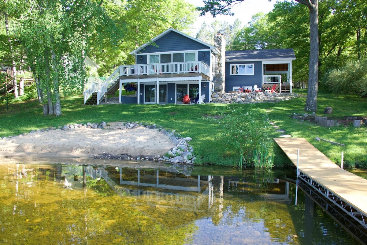 A two-story lakefront property features a gray exterior with a charming stone fireplace. A spacious deck with white railings overlooks the water, reflecting the surrounding green trees. A sandy beach area is visible in the foreground, with a wooden dock extending into the lake.