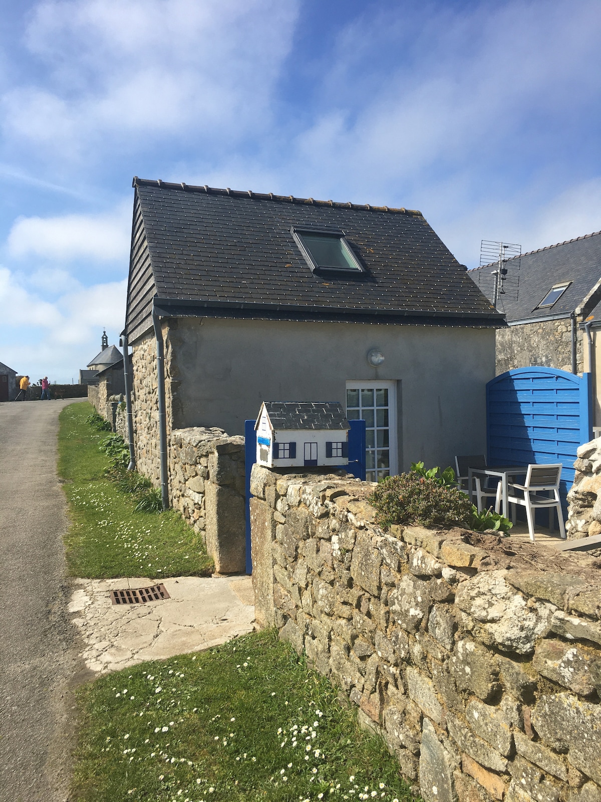 A quaint independent house is shown with a slate roof and stone exterior. A small outdoor area features a table and chairs beside a blue structure. Nearby greenery adds to the calm atmosphere, with clouds partially obscuring a bright blue sky.