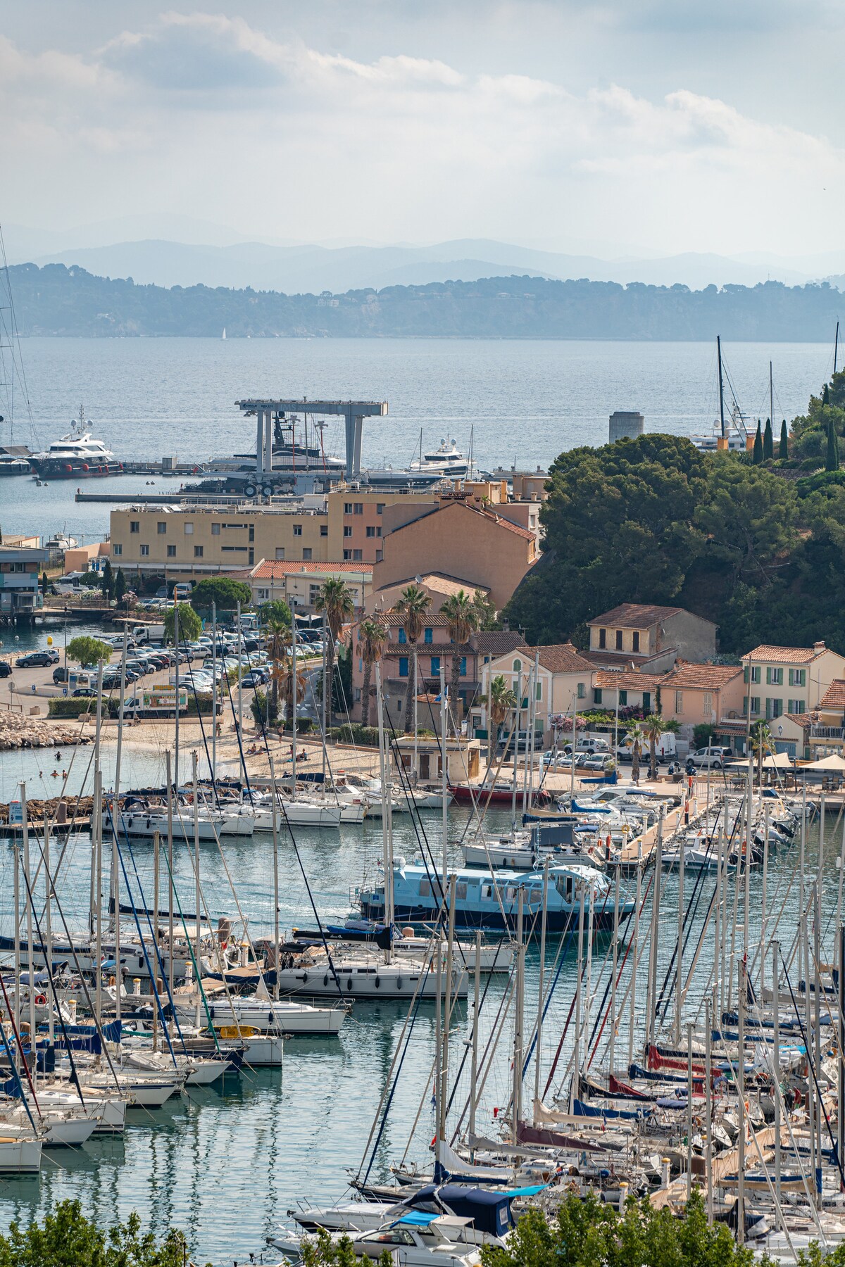 A view of the marina reveals numerous sailboats and yachts moored along the docks. The picturesque shoreline is lined with varied architecture, including pastel-colored buildings. The backdrop features distant hills under a partly cloudy sky, providing a serene seaside atmosphere.