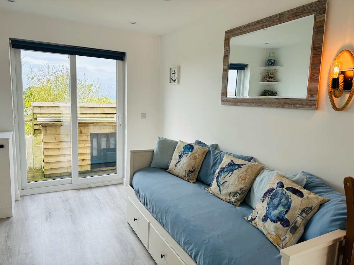 A bright seating area features a large blue sofa adorned with decorative pillows. Natural light floods through sliding glass doors, which provide access to an outdoor deck. A mirror and wall sconce enhance the ambiance, while a shelf with decor items adds a touch of detail.