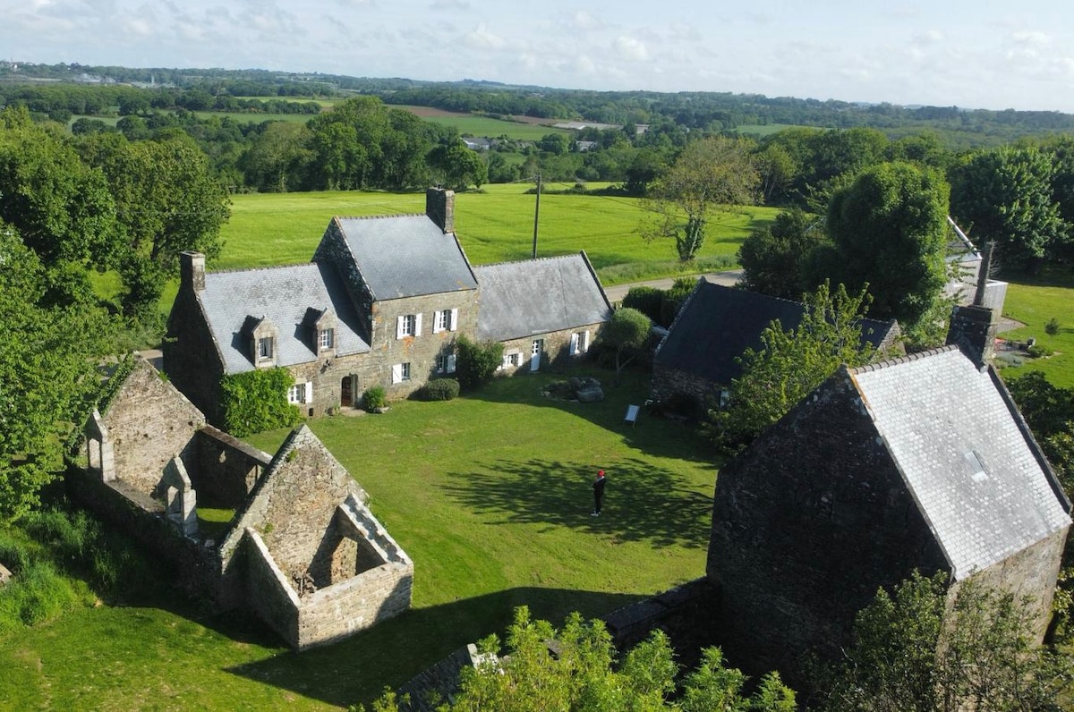An aerial view showcases a cluster of rustic stone buildings arranged around a green courtyard, surrounded by lush fields. Large trees provide shade, and the landscape extends into the distance under a clear sky, enhancing the serene rural setting.