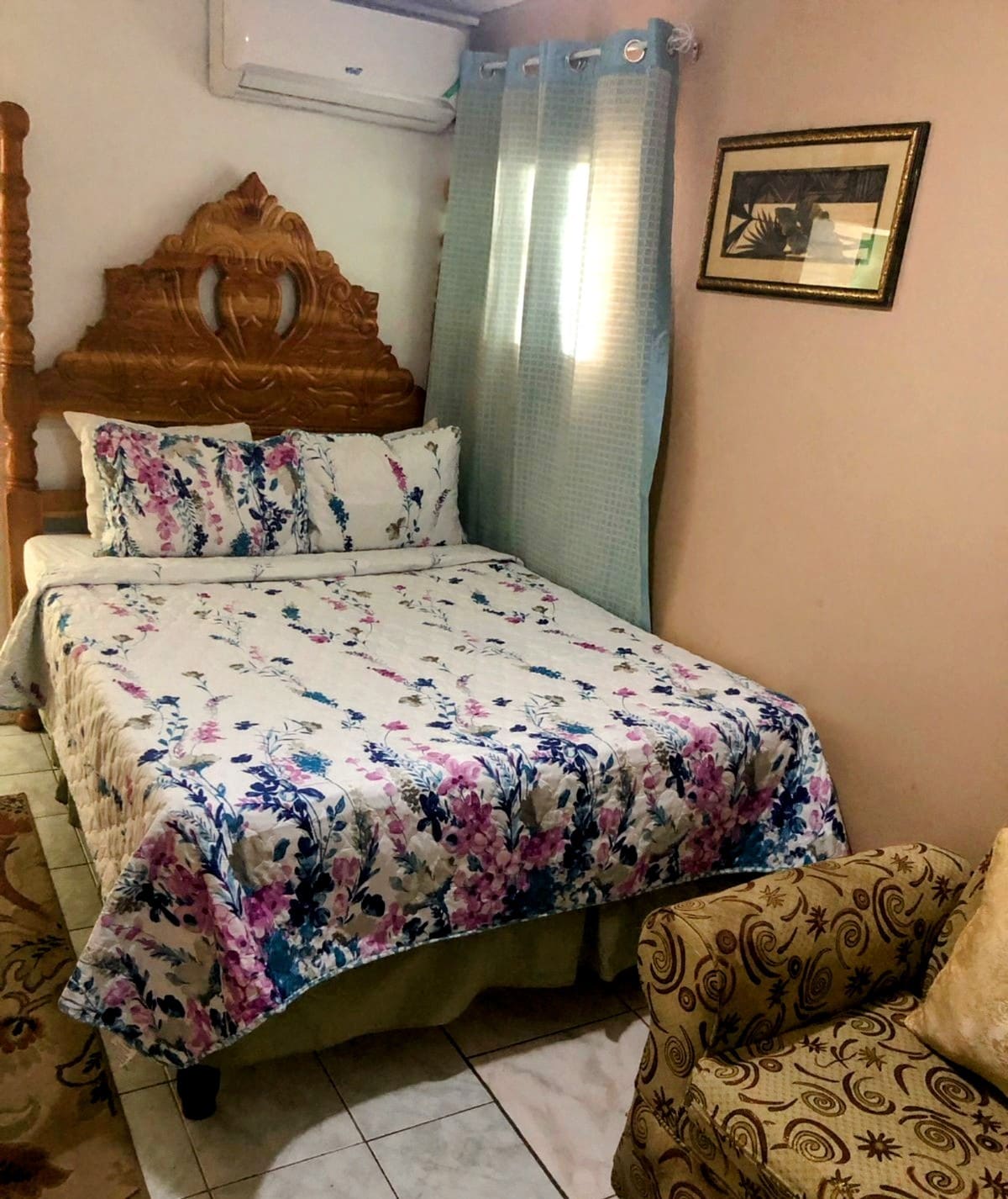 A cozy bedroom features a wooden bed with a floral-patterned bedspread. A small armchair in a paisley design complements the space. Natural light enters through two windows adorned with light blue curtains, creating a bright and comfortable atmosphere.