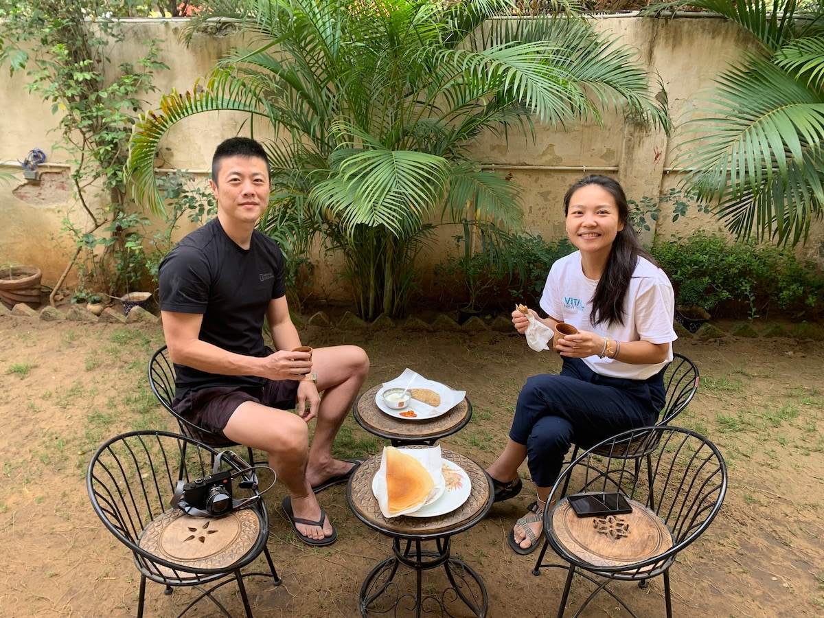 A shaded outdoor seating area features two individuals seated at a small black table. Two round chairs with intricate designs complement the table, which holds a plate of food and beverages. Lush greenery surrounds the space, adding to a relaxed atmosphere.