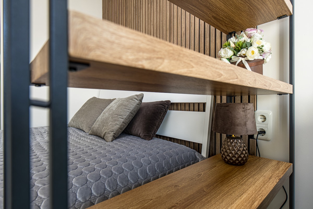A close-up of a wooden shelving unit displaying a small flower arrangement in a pot. Below, a plush bed with textured gray bedding features decorative pillows. A bedside lamp emits soft light beside a power outlet, providing functional elegance.