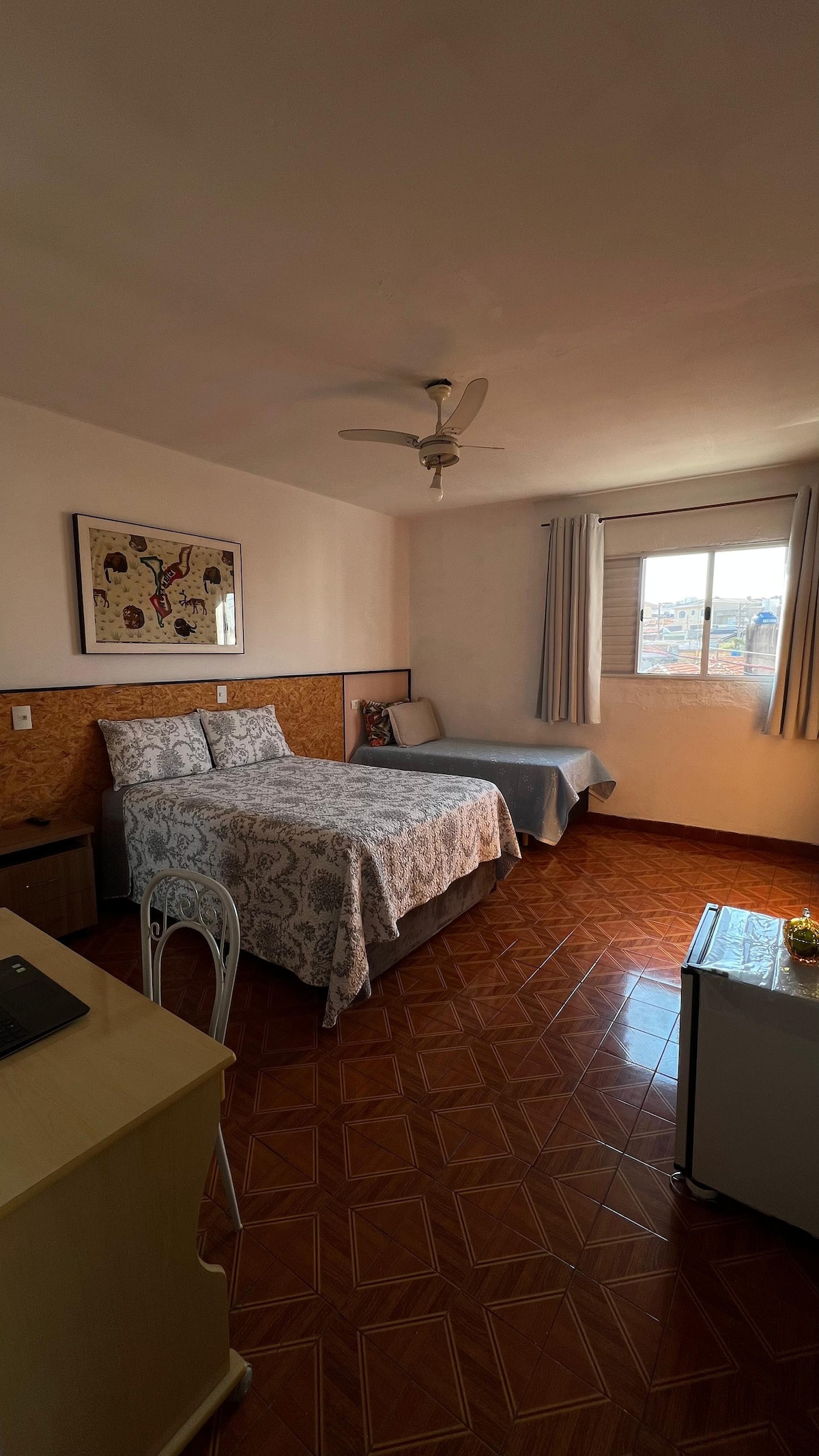 A spacious guest room is visible, featuring a double bed with patterned bedding and a single bed in a light color. A window allows natural light to fill the space, and a small desk is positioned nearby. A fan is mounted on the ceiling.