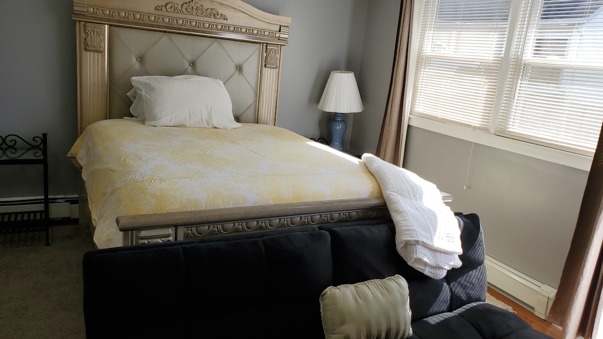 A large bed with an intricately designed headboard is positioned against a grey wall. A pair of windows dressed with sheer blinds allow natural light to brighten the space. A dark sofa is situated nearby, with a folded blanket resting on the arm.