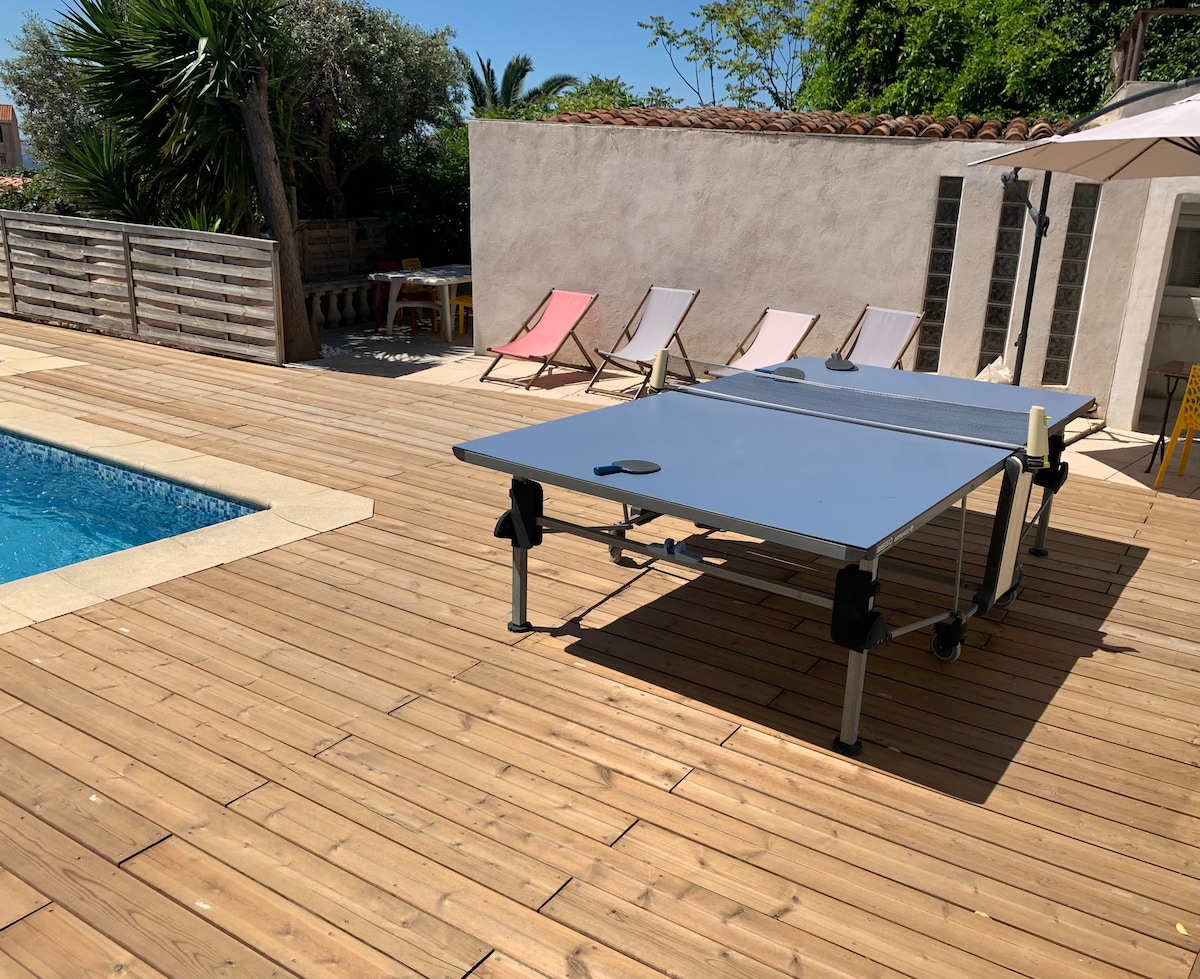 An outdoor area is shown, featuring a wooden deck alongside a swimming pool. A table tennis table is centrally located, complemented by several lounge chairs in various pastel colors. Lush greenery and a wall are visible in the background, providing a serene environment.