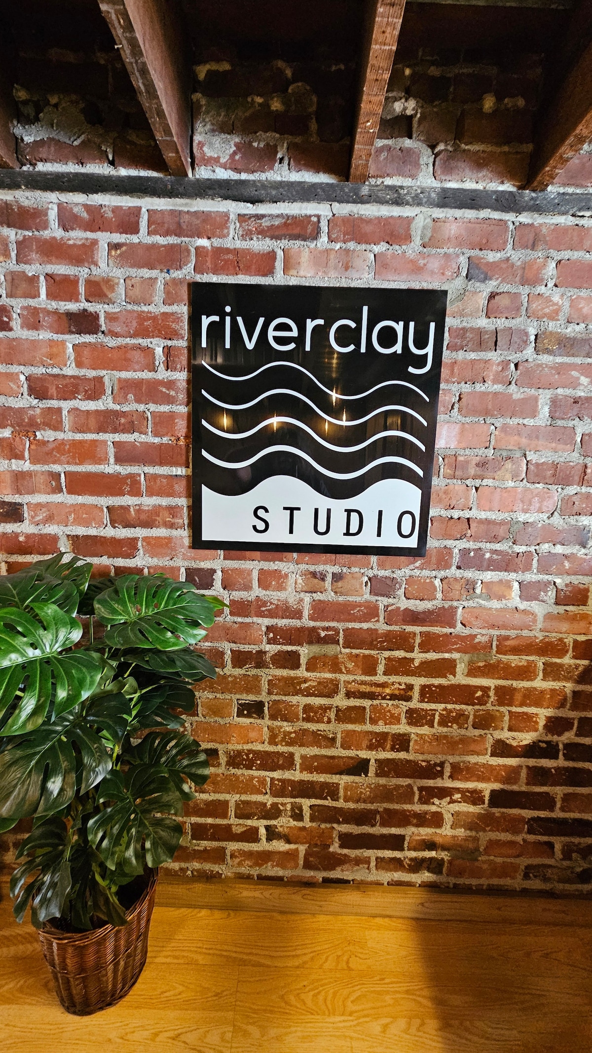 A wooden sign displaying the text 'riverclay STUDIO' is mounted on a brick wall. The design features flowing lines, creating a sense of movement. A potted plant with broad green leaves is placed nearby on a wooden floor, enhancing the studio's welcoming ambiance.