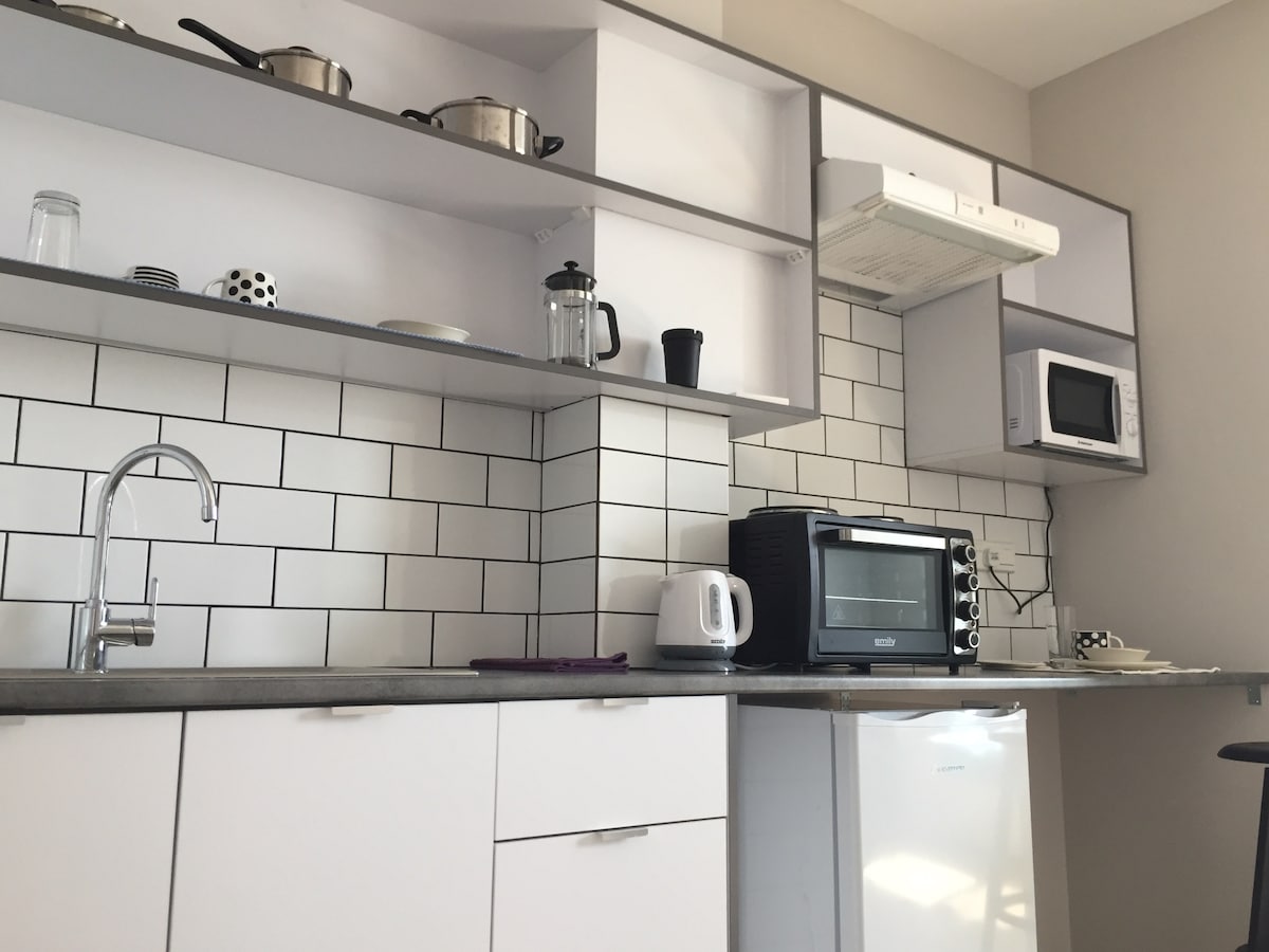 A functional kitchen area displays modern appliances, including a microwave, kettle, and coffee maker. Open shelving holds cookware and utensils, while white cabinetry contrasts with the dark countertop. Tiled backsplash adds a clean and contemporary feel to the space.