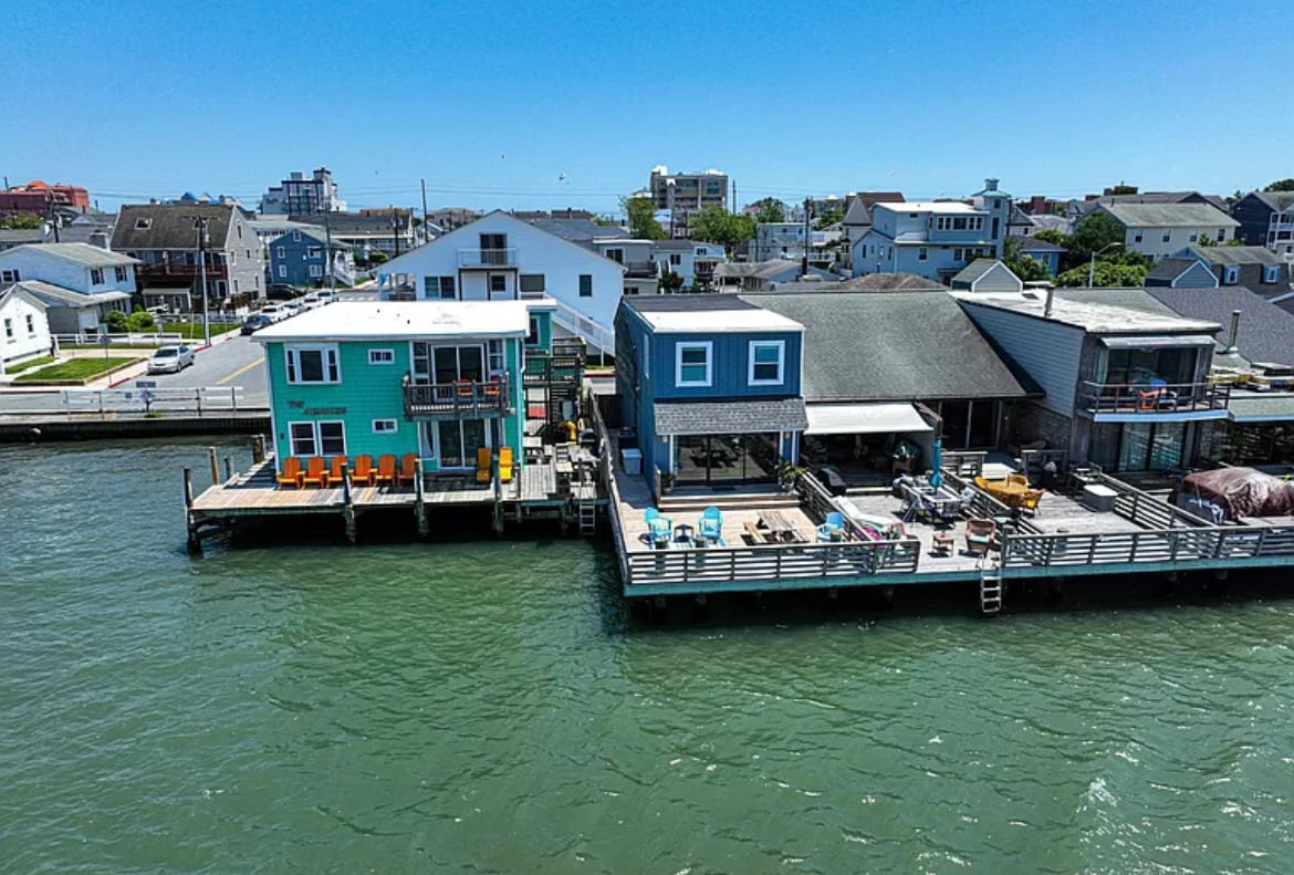 Aerial view of waterfront homes in Ocean City, showcasing two distinct residences. One features vibrant teal exterior and several bright orange lounge chairs on the deck. The other, with a more subdued design, has various outdoor seating areas and a dock extending into the water.