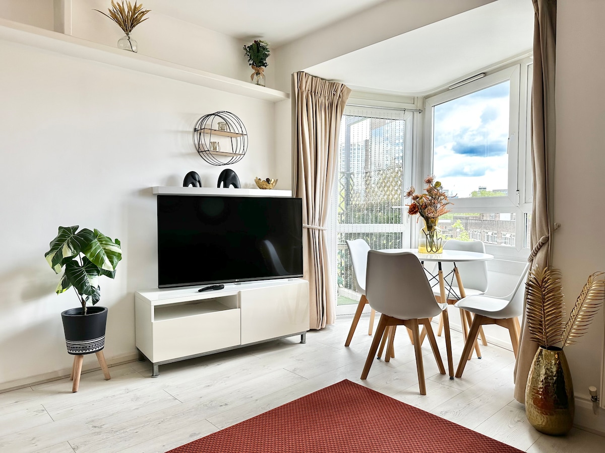 A bright living area showcases a modern design, featuring a large television on a white cabinet. A round dining table with chairs is positioned near the window, complemented by indoor plants and decorative accents. Natural light fills the space, enhancing the airy atmosphere.